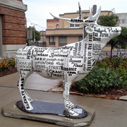 White cow sculpture covered in printed words, standing on a sidewalk in front of brick buildings and flowers