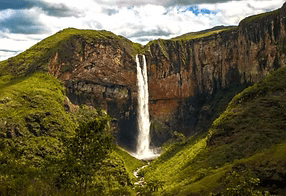 Alta cachoeira despencando entre penhascos verdes em um exuberante vale montanhoso sob um céu nublado.