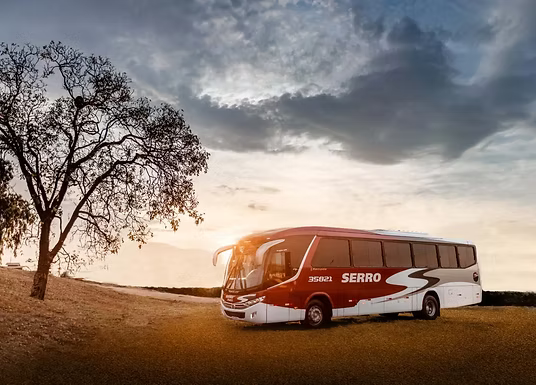 Ônibus de passageiros vermelho e branco trafegando por uma estrada rural ao pôr do sol, ao lado de uma árvore sem folhas.