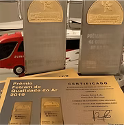 Troféus e um certificado de premiação de qualidade do ar de 2019 em exposição, com um veículo vermelho ao fundo.
