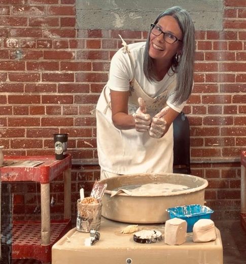 Tracy DiBease standing over a pottery wheel giving the thumbs up sign