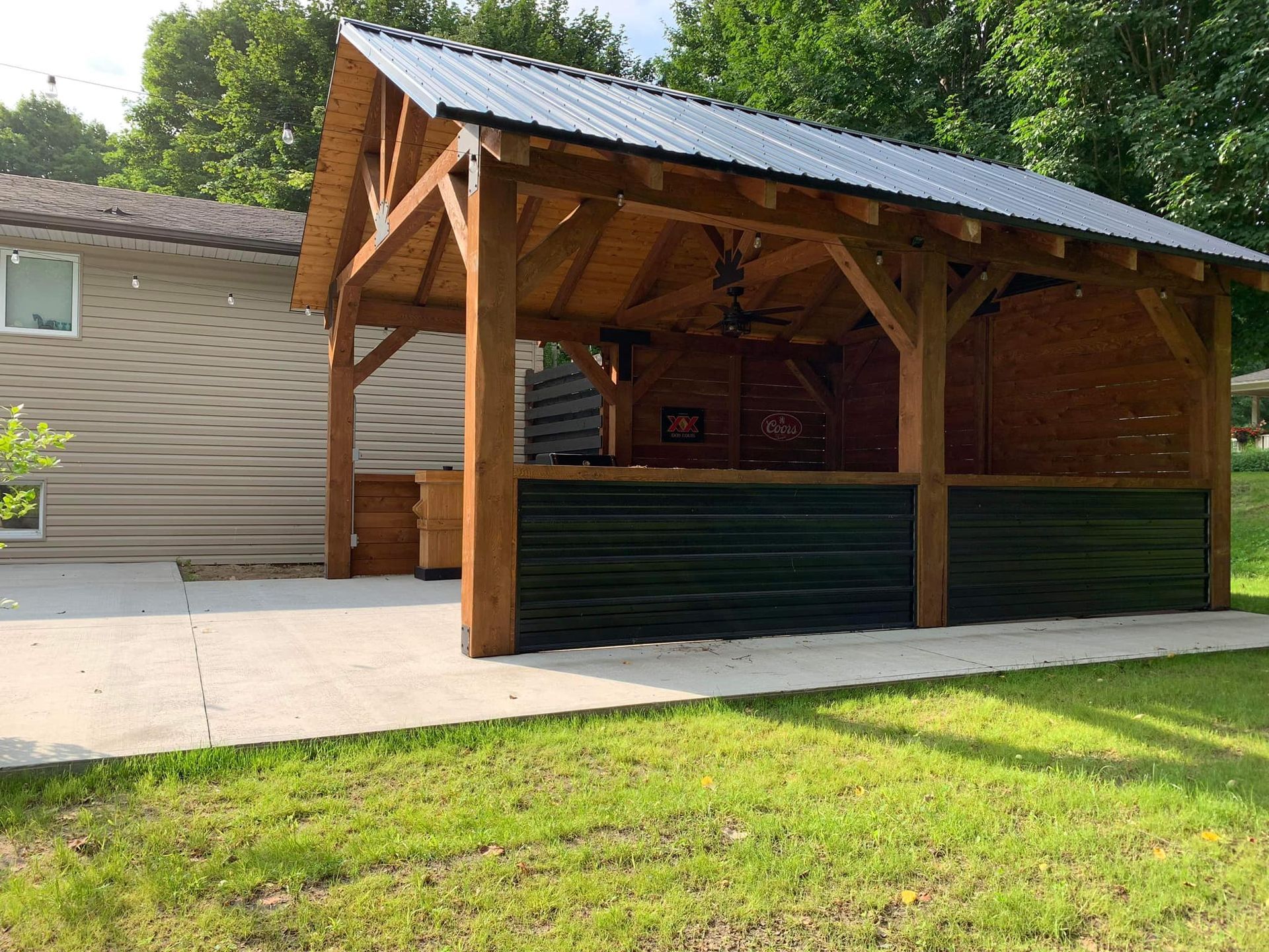 Pavillon en bois avec un toit en métal, situé à côté d'un bâtiment, sur une pelouse.