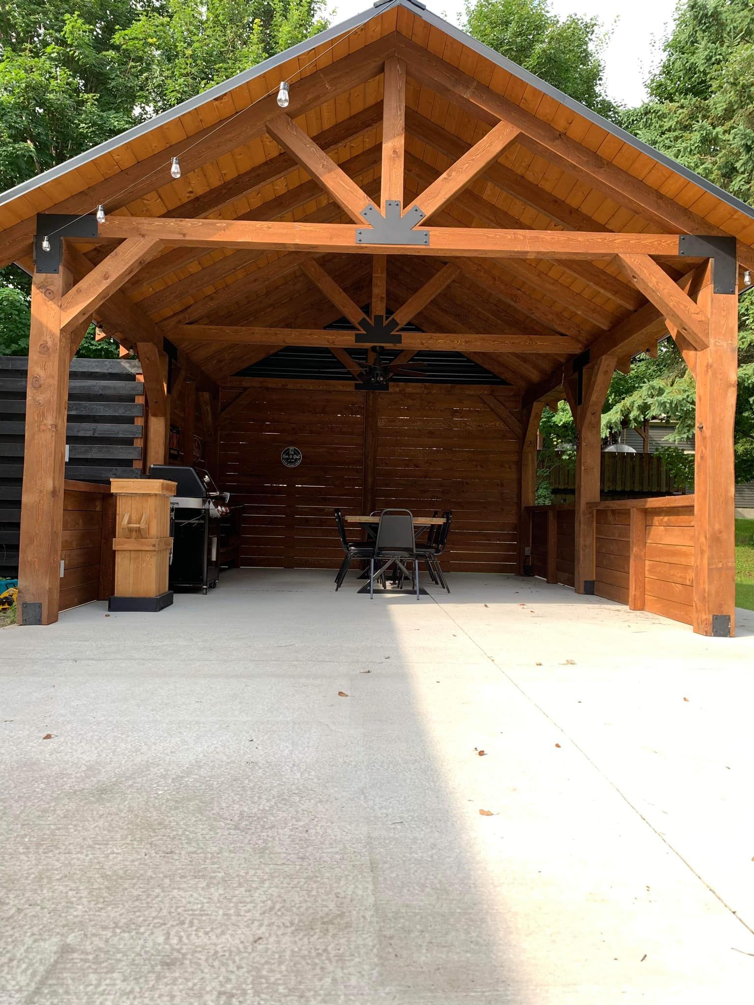 Pavillon en bois à ciel ouvert avec sol en béton, tables, chaises et ventilateur de plafond sous un toit pointu.