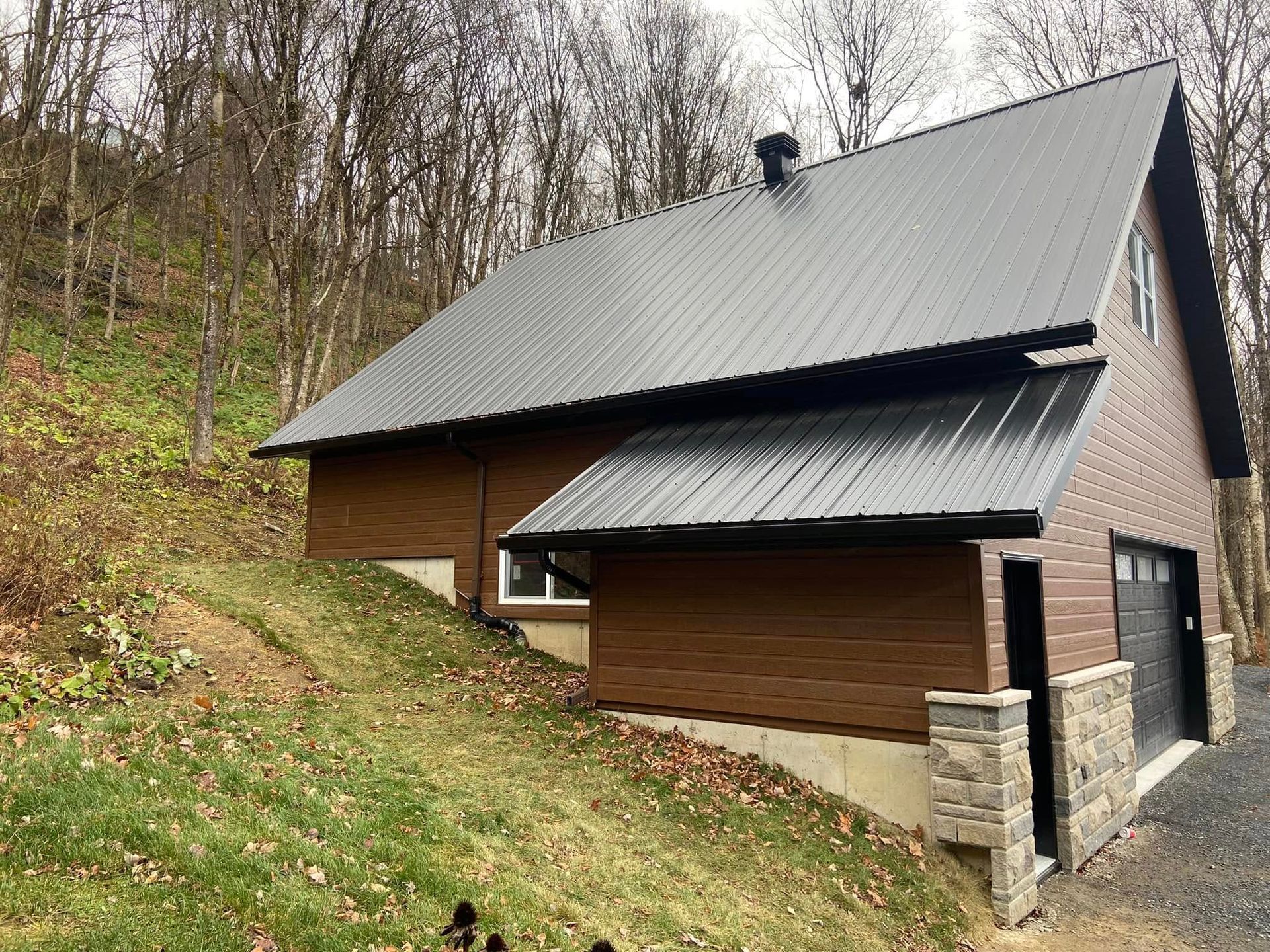 Petite maison brune au toit gris pentu, sur un flanc de colline boisé.
