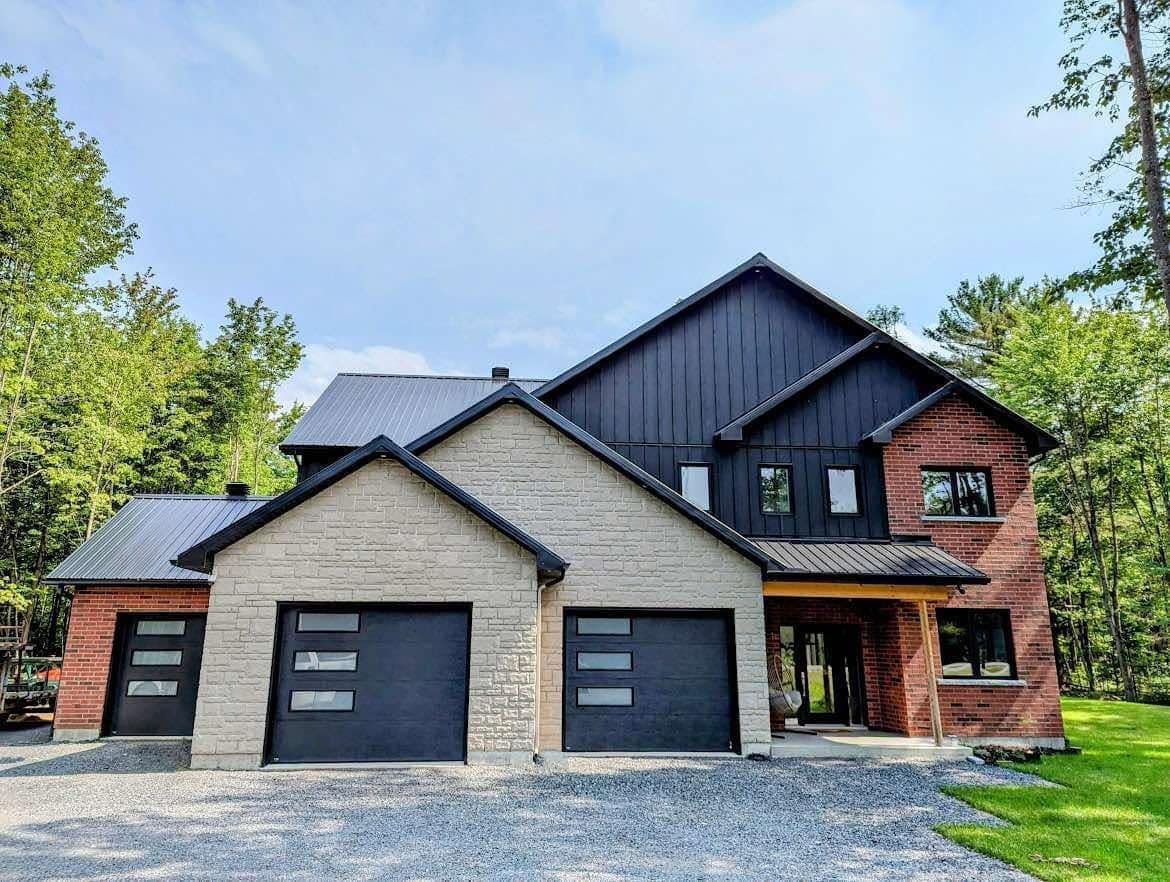 Maison moderne à deux étages avec portes de garage grises, façade en briques et en bois foncé, entourée d'arbres