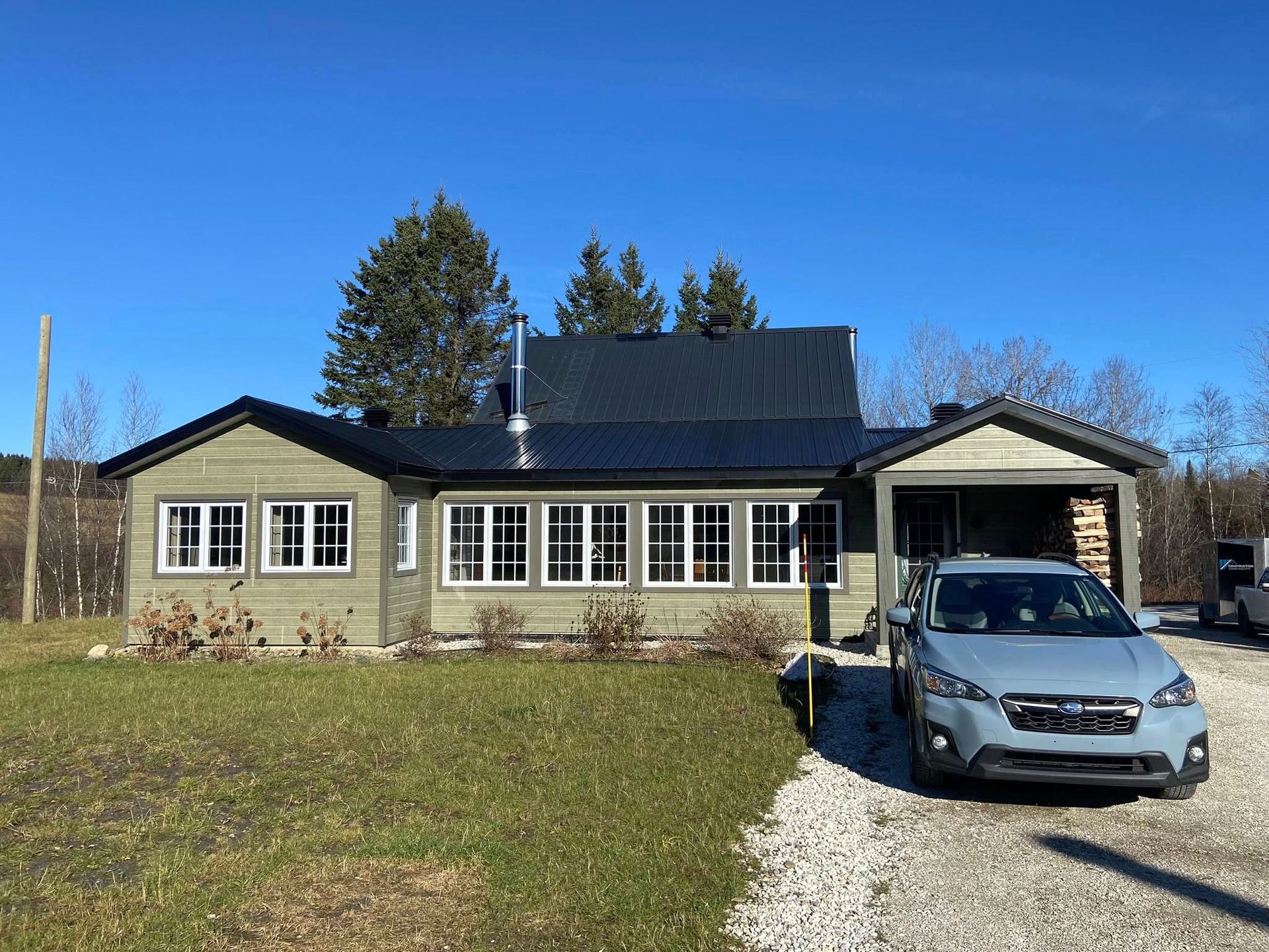 Maison de plain-pied au toit sombre et voiture bleue garée sur une allée de gravier sous un ciel dégagé.