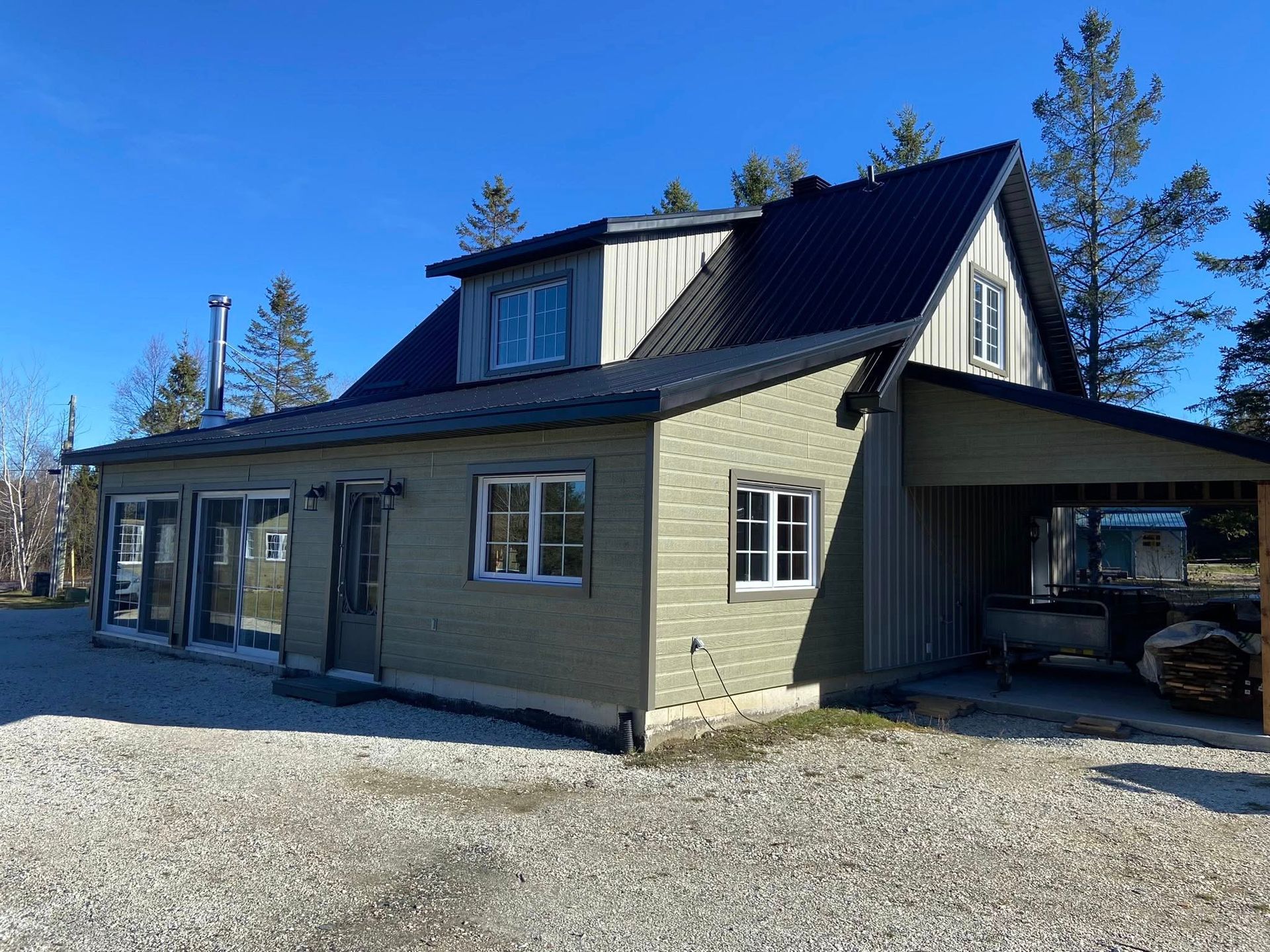 Maison grise avec toit noir et abri voiture sur un terrain ensoleillé recouvert de gravier.