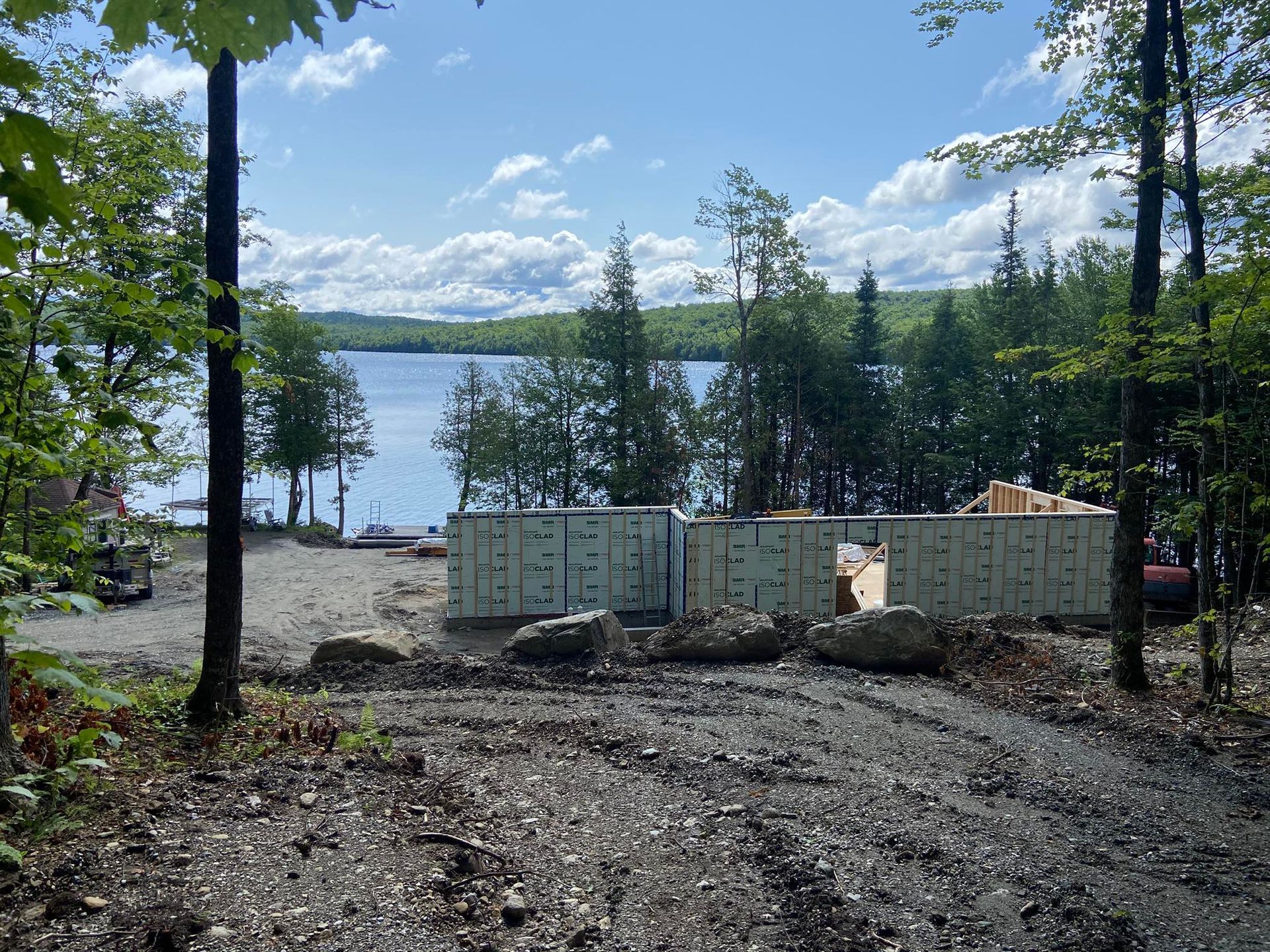Terrain vague défriché avec des conteneurs maritimes empilés au bord d'un lac boisé sous un ciel d'un bleu éclatant