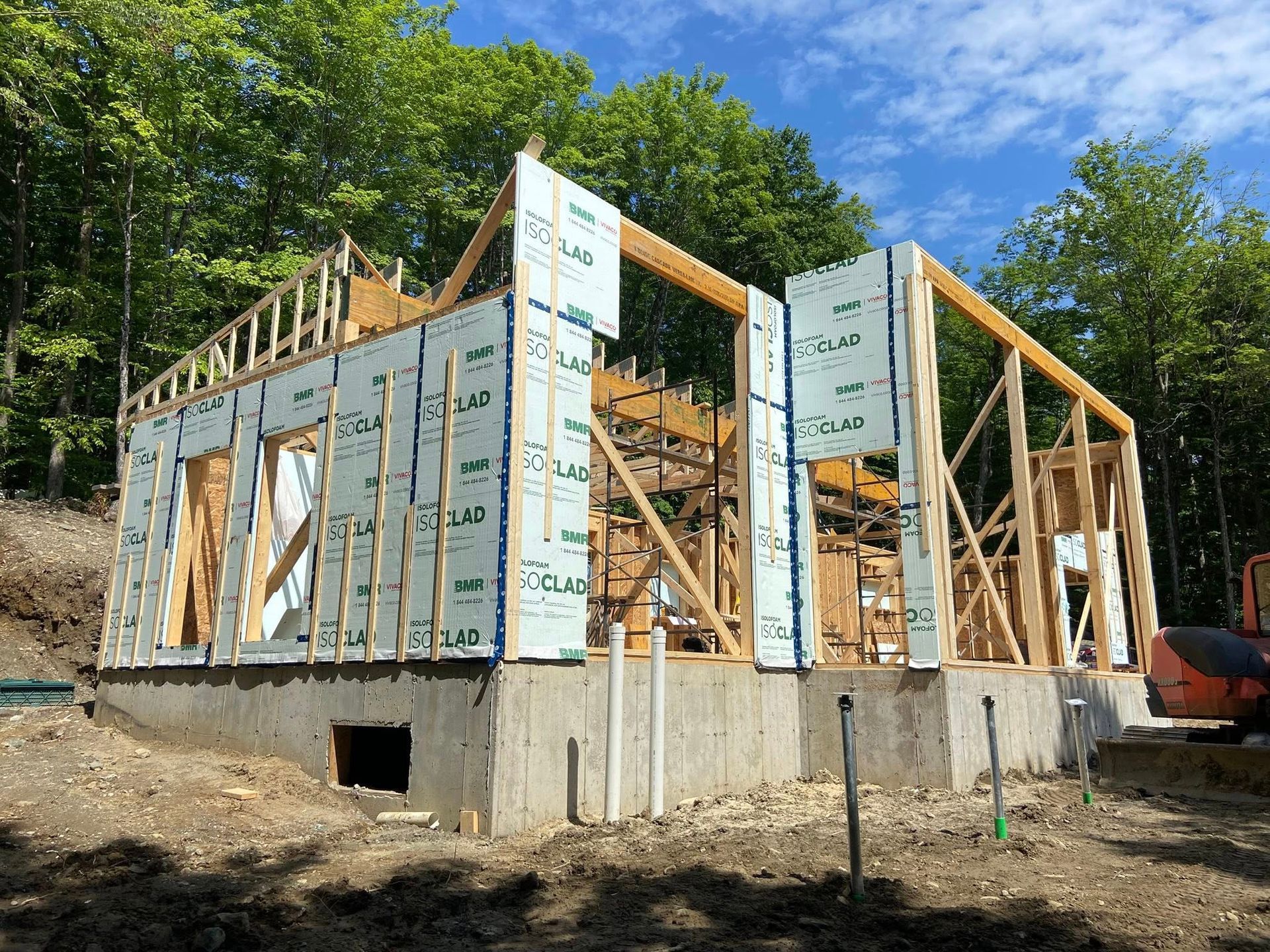 Maison partiellement à ossature bois en construction, avec panneaux isolants bleus et fondations en béton sur un terrain boisé