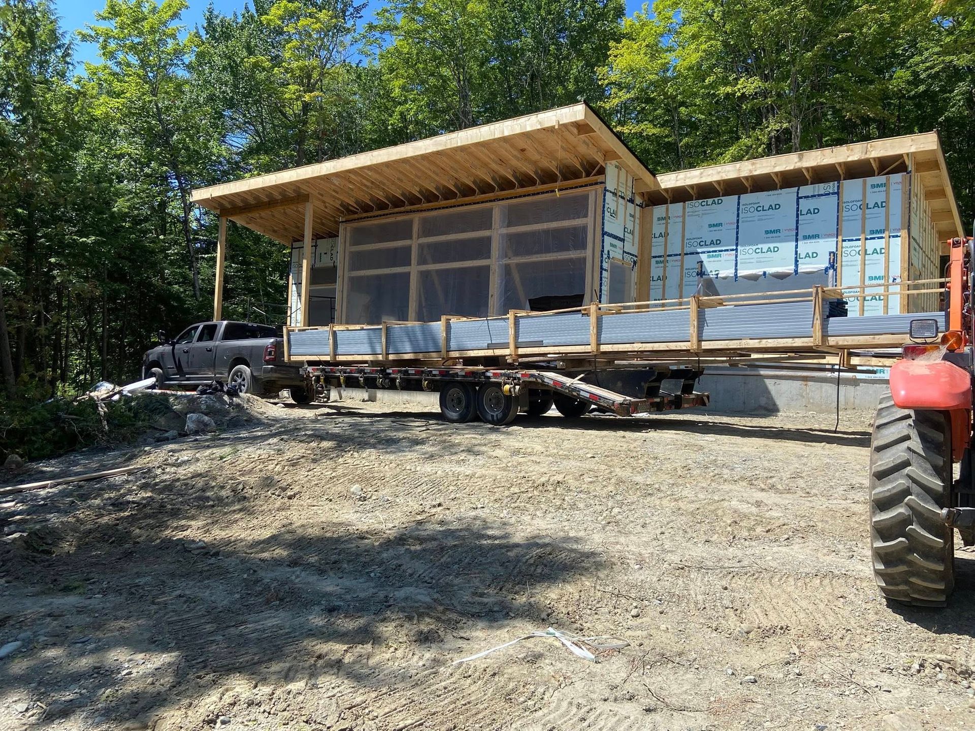 Une mini-maison modulaire en construction sur une remorque dans une clairière boisée, avec un tracteur à proximité.