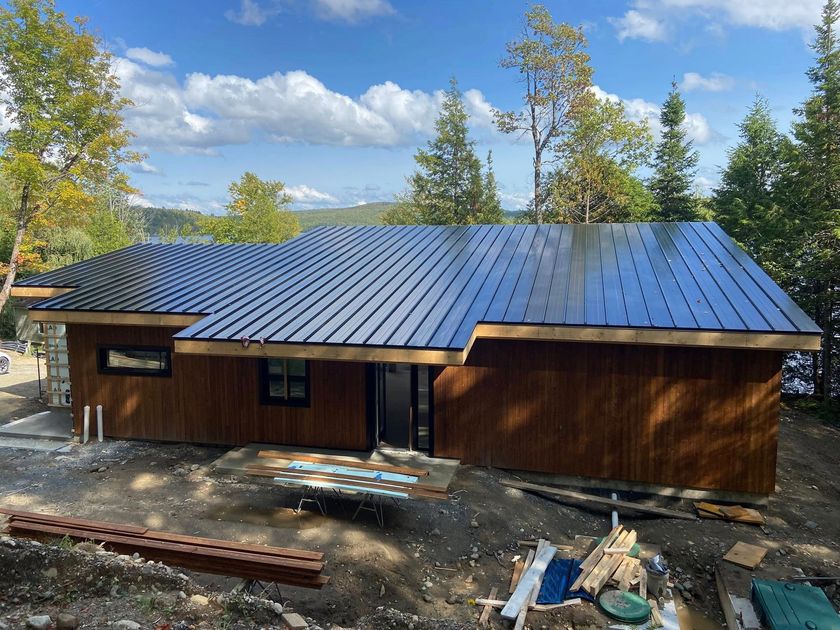 Maison en bois de plain-pied avec un toit en métal sombre, entourée d'arbres sous un ciel bleu.