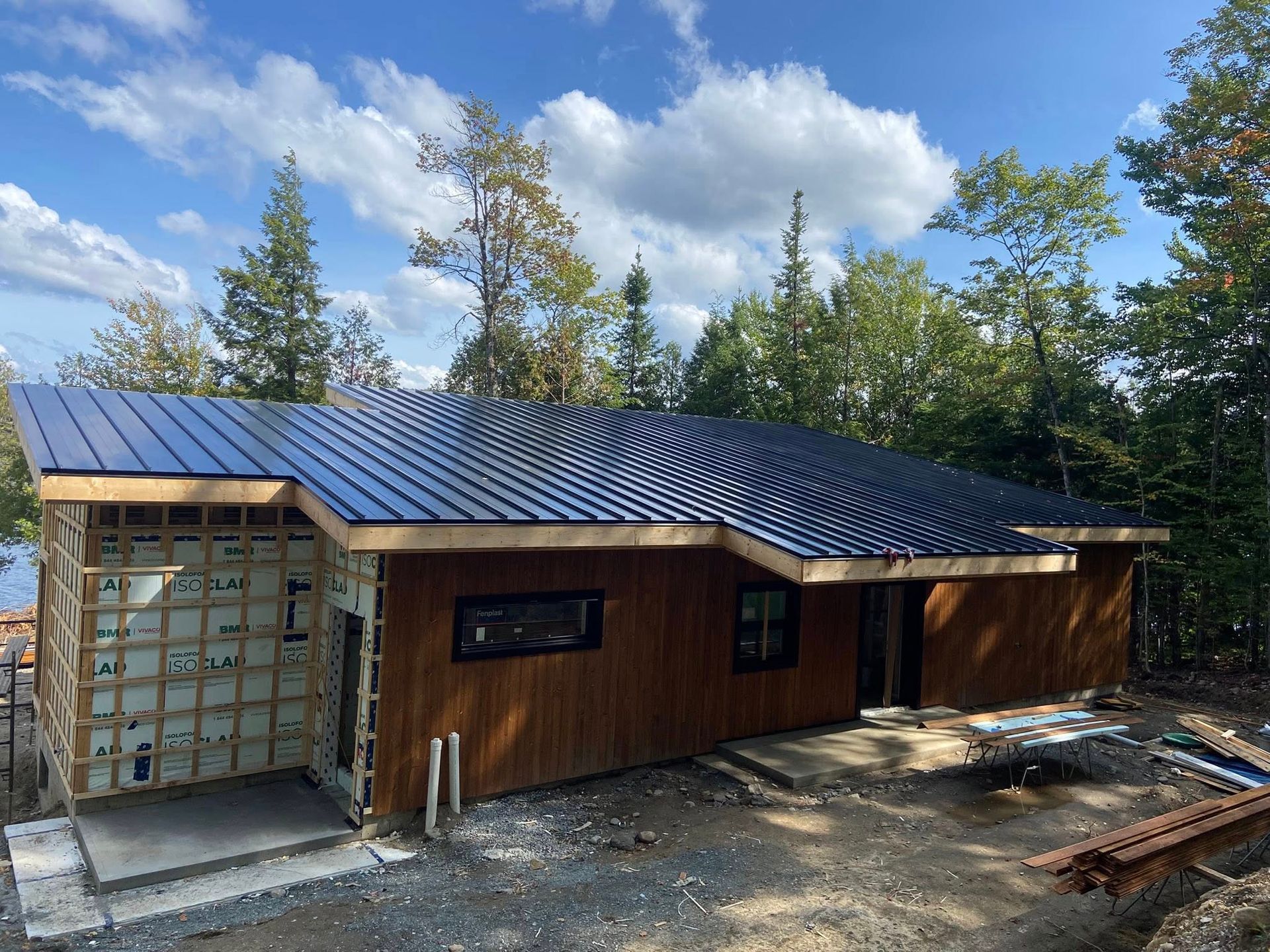 Petite maison en bois en construction, avec un toit en métal bleu, des fenêtres et des arbres tout autour, sous un ciel lumineux.