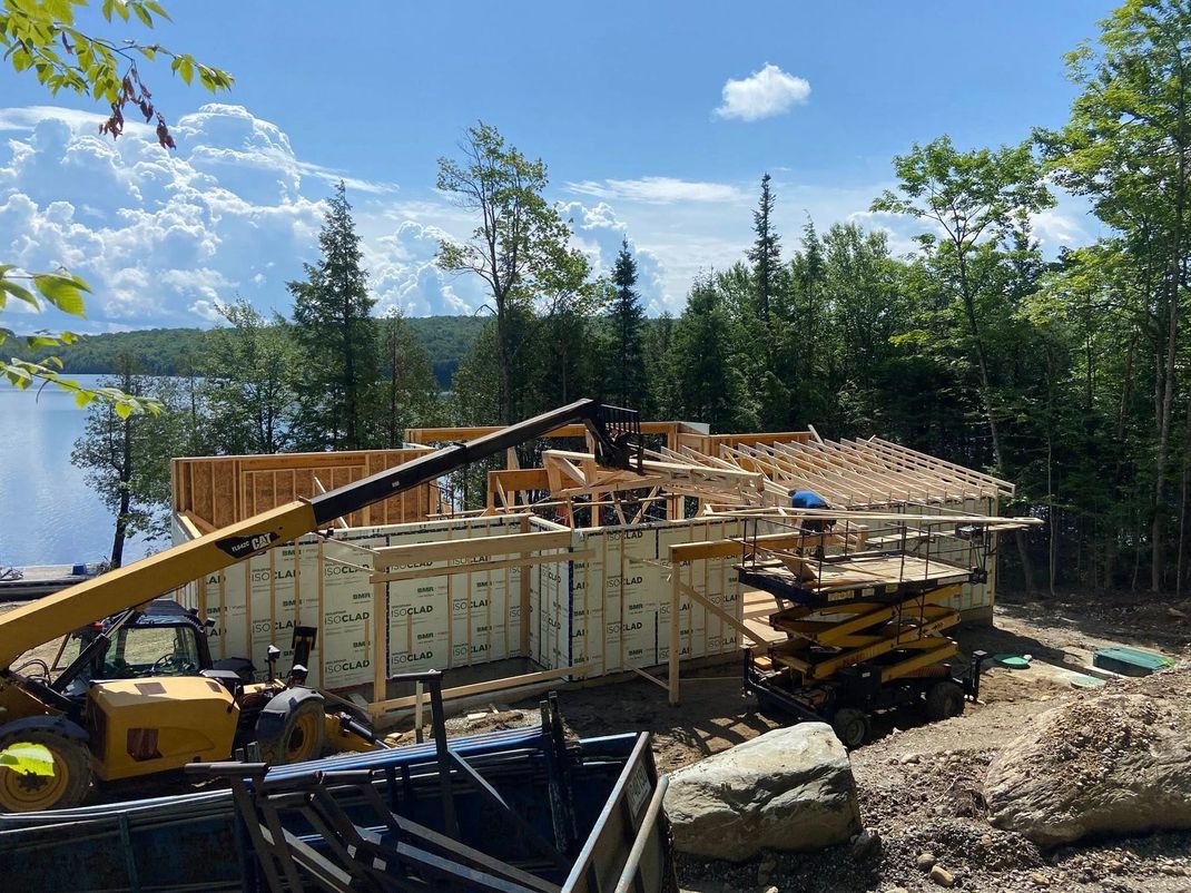 Chantier de construction au bord d'un lac, avec une maison partiellement charpentée, une grue, du bois et des arbres sous un ciel bleu