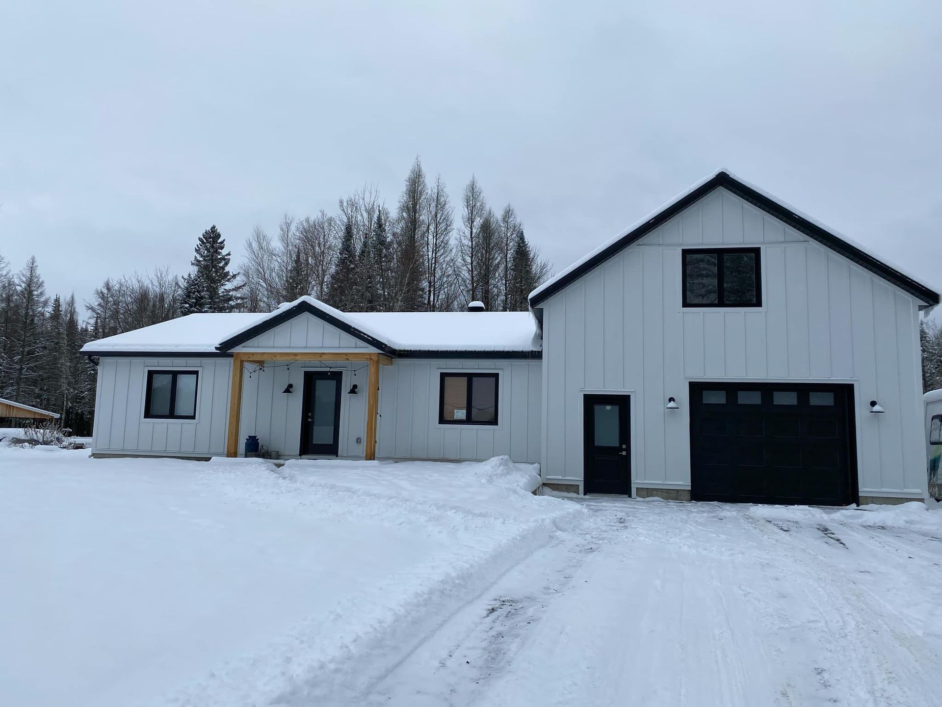 Maison et garage enneigés, avec allée, se détachant sur un fond d'arbres dénudés en hiver.