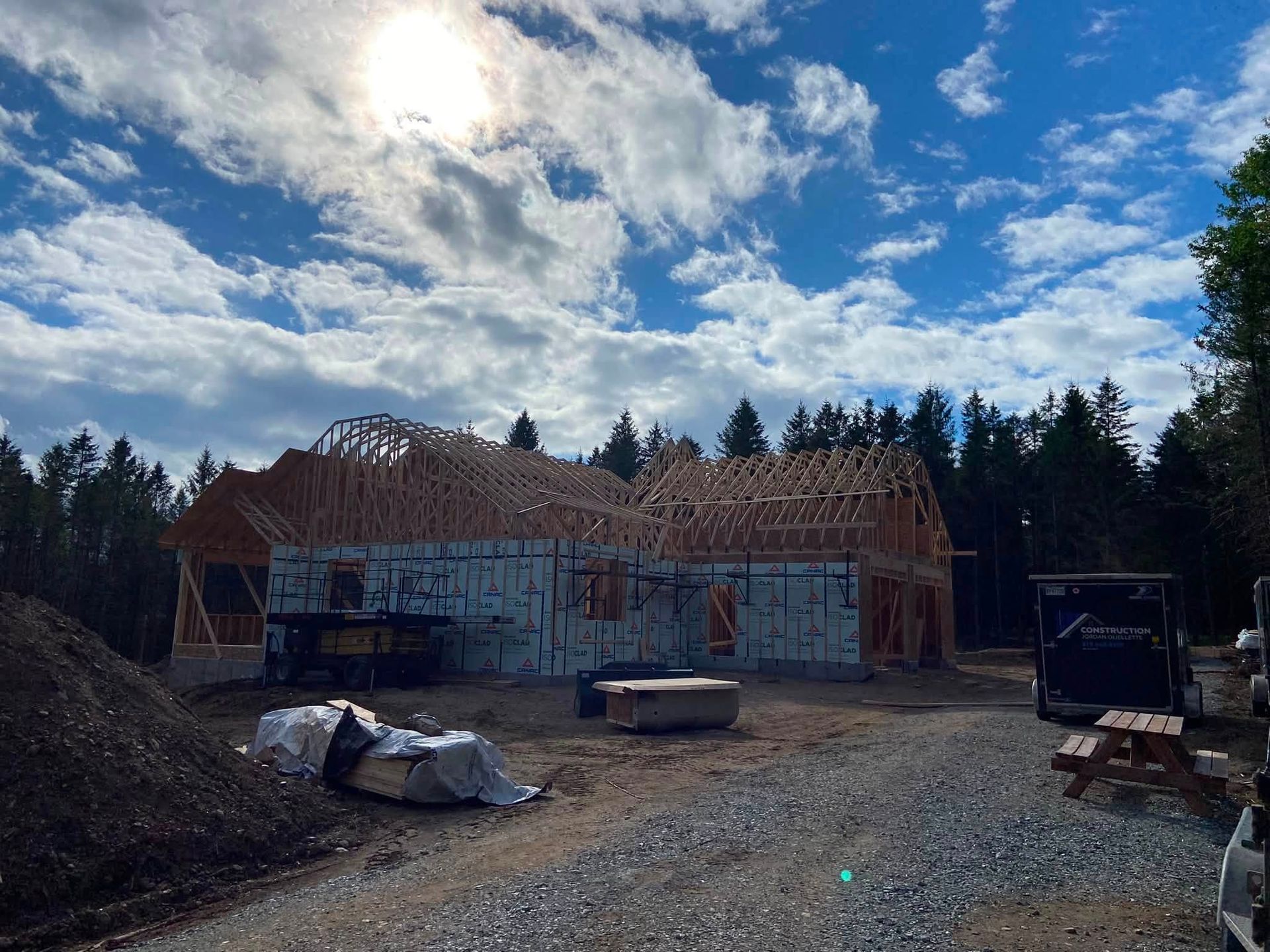 Maison en construction, à la charpente apparente, sous un ciel nuageux, entourée de matériaux de construction et d'arbres.