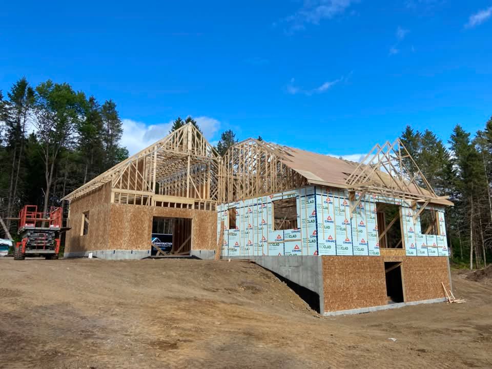 Maison partiellement construite avec charpente apparente et murs en contreplaqué sur un terrain en terre battue sous un ciel bleu