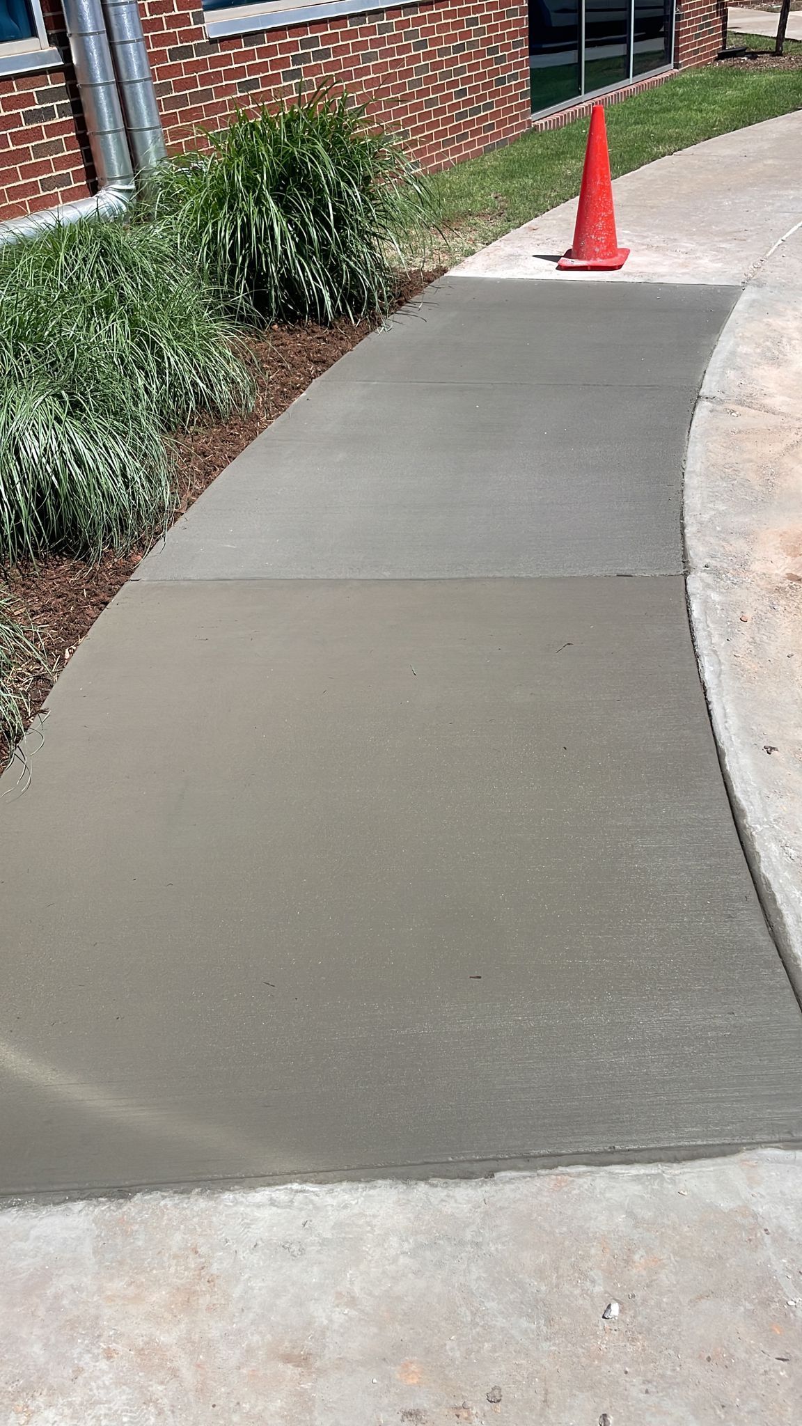 Concrete sidewalk with a curved edge, freshly poured, orange traffic cone in the distance.