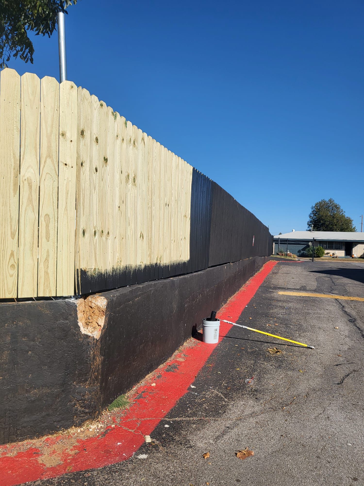 A wooden fence partially painted black alongside a parking lot, a paint bucket, and a paint roller nearby.