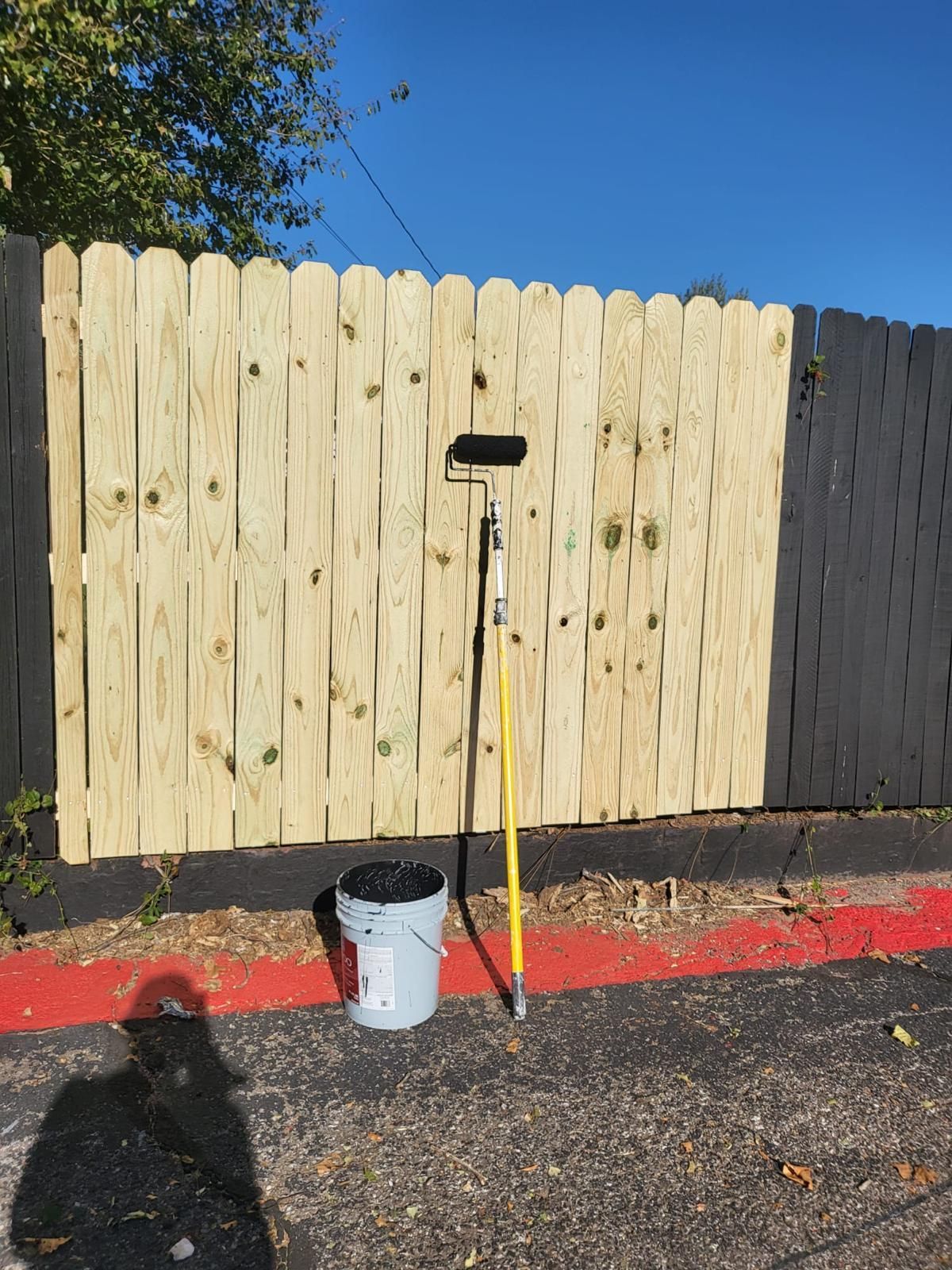 Wooden fence being painted; half natural wood, half black. Paint roller and bucket sit in front.