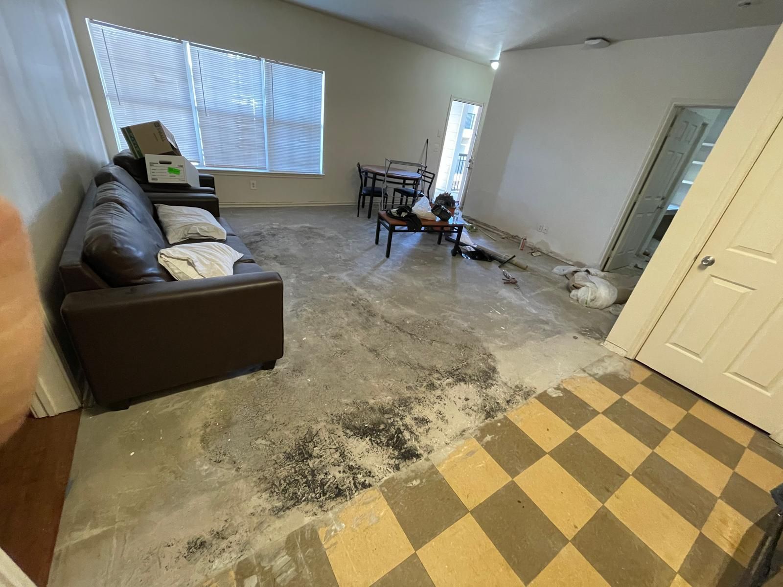 Living room with brown sofa, damaged floor, checkered tile, open door, and table.