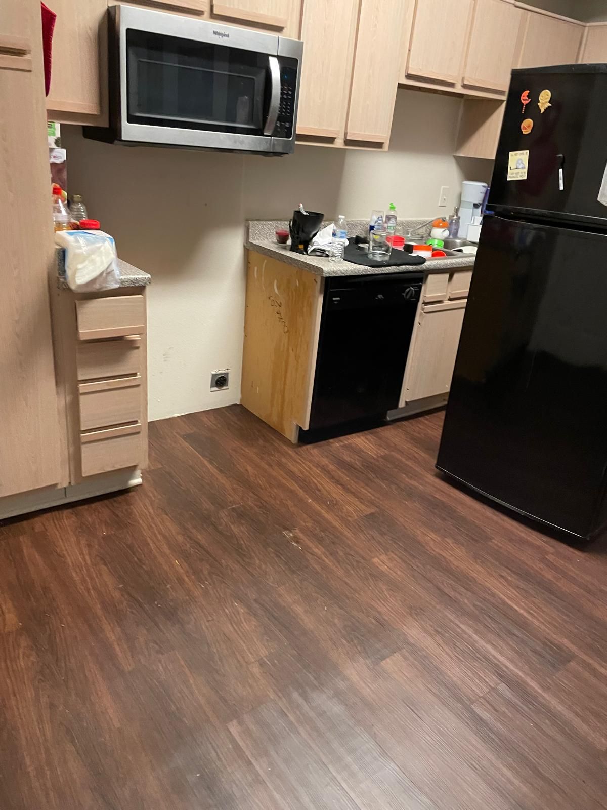 A kitchen with wood flooring, cabinets, a microwave, black appliances, and countertops with items on them.