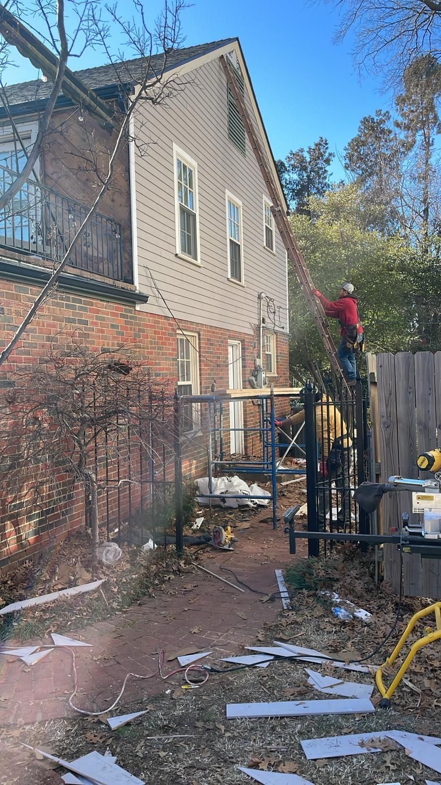 Construction worker on a ladder beside a two-story house, brick and gray siding. Fencing and debris in the yard.