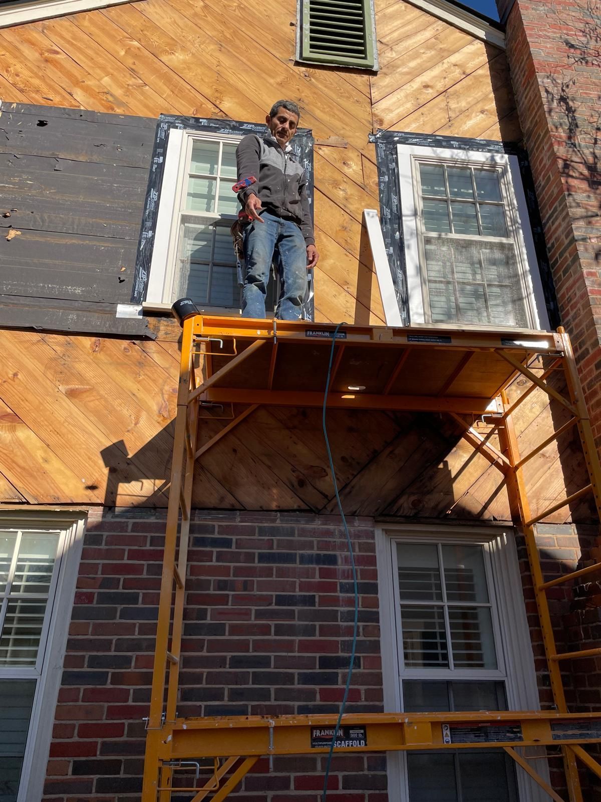 Person on scaffolding working on exterior wall. Brown siding, brick, windows.