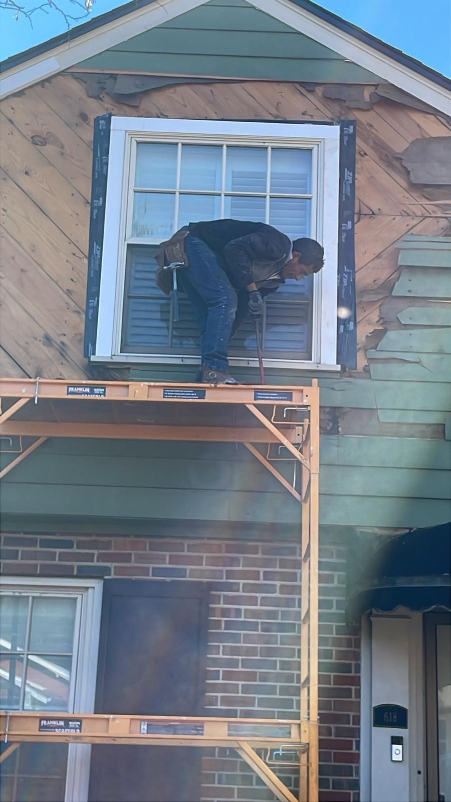 Person working on house siding, standing on scaffolding. Exterior has peeling paint, blue and brown tones.
