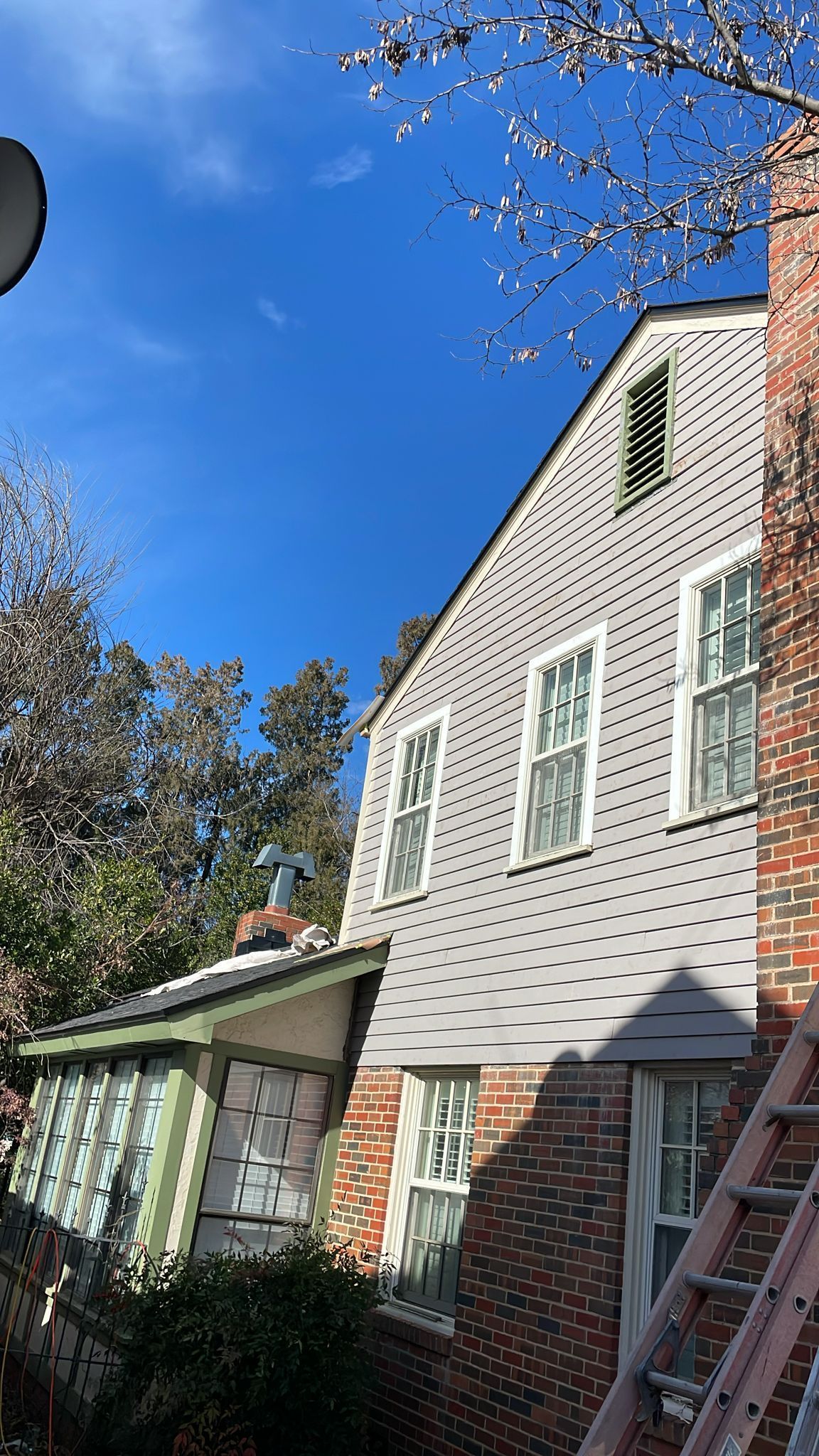 Side view of a house with gray siding, brick chimney, and a ladder against the wall under a bright blue sky.