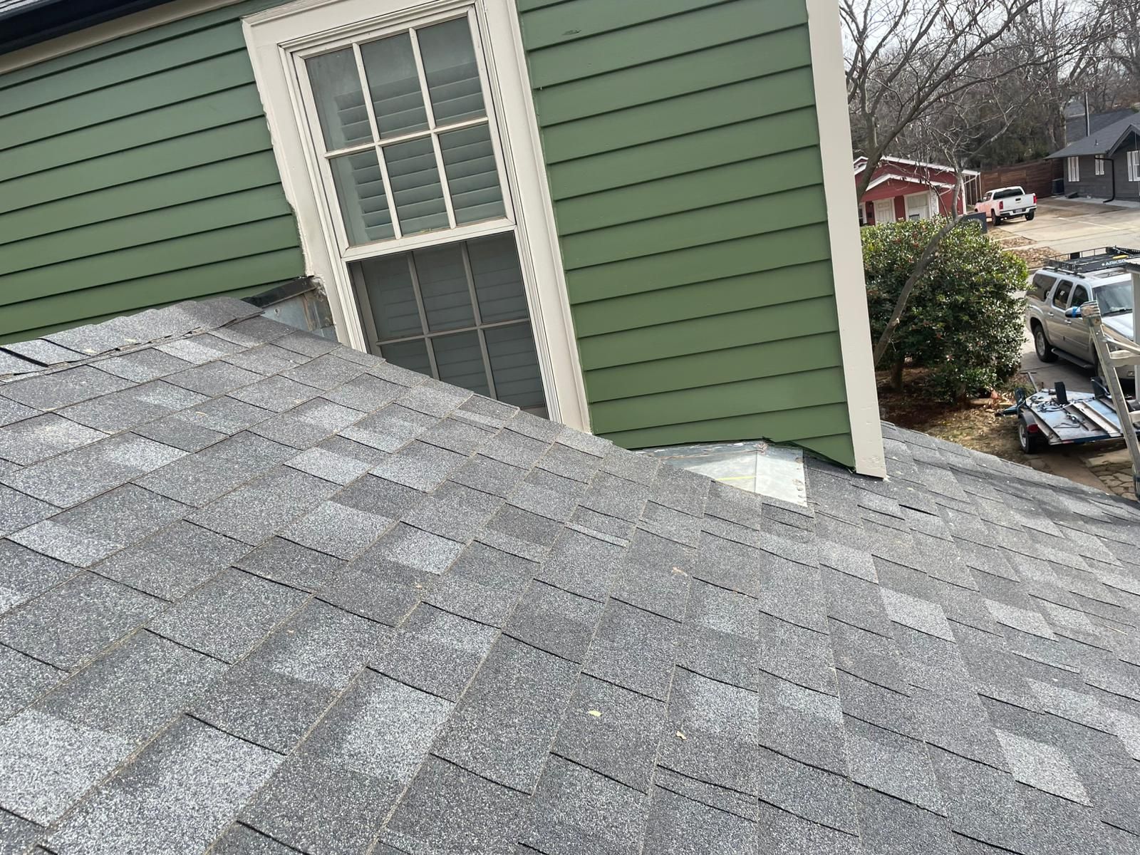 Close-up of a gray shingle roof next to a green-sided house with a white-framed window.