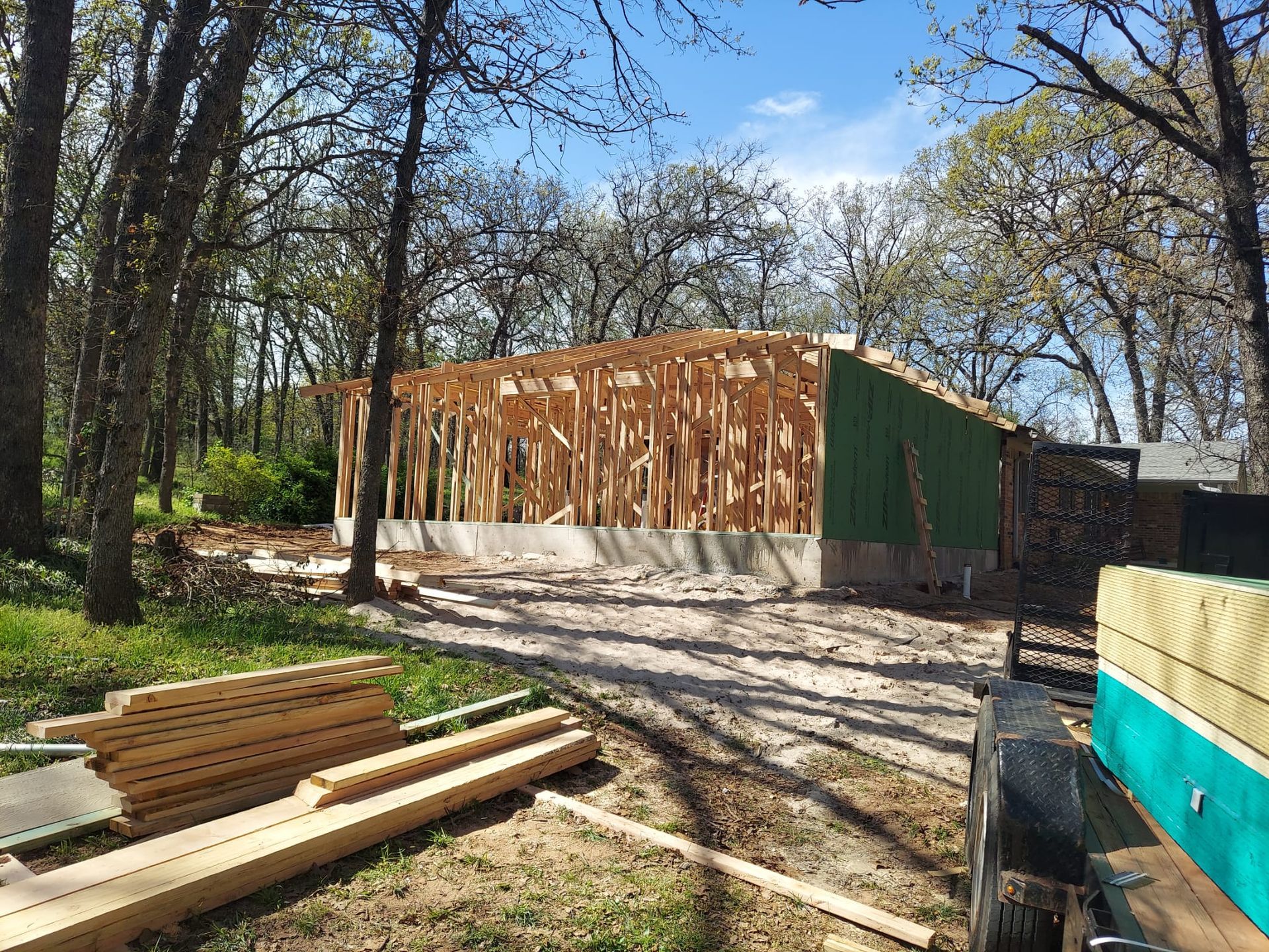 Construction of a wood-framed building in a wooded area; green sheathing visible.