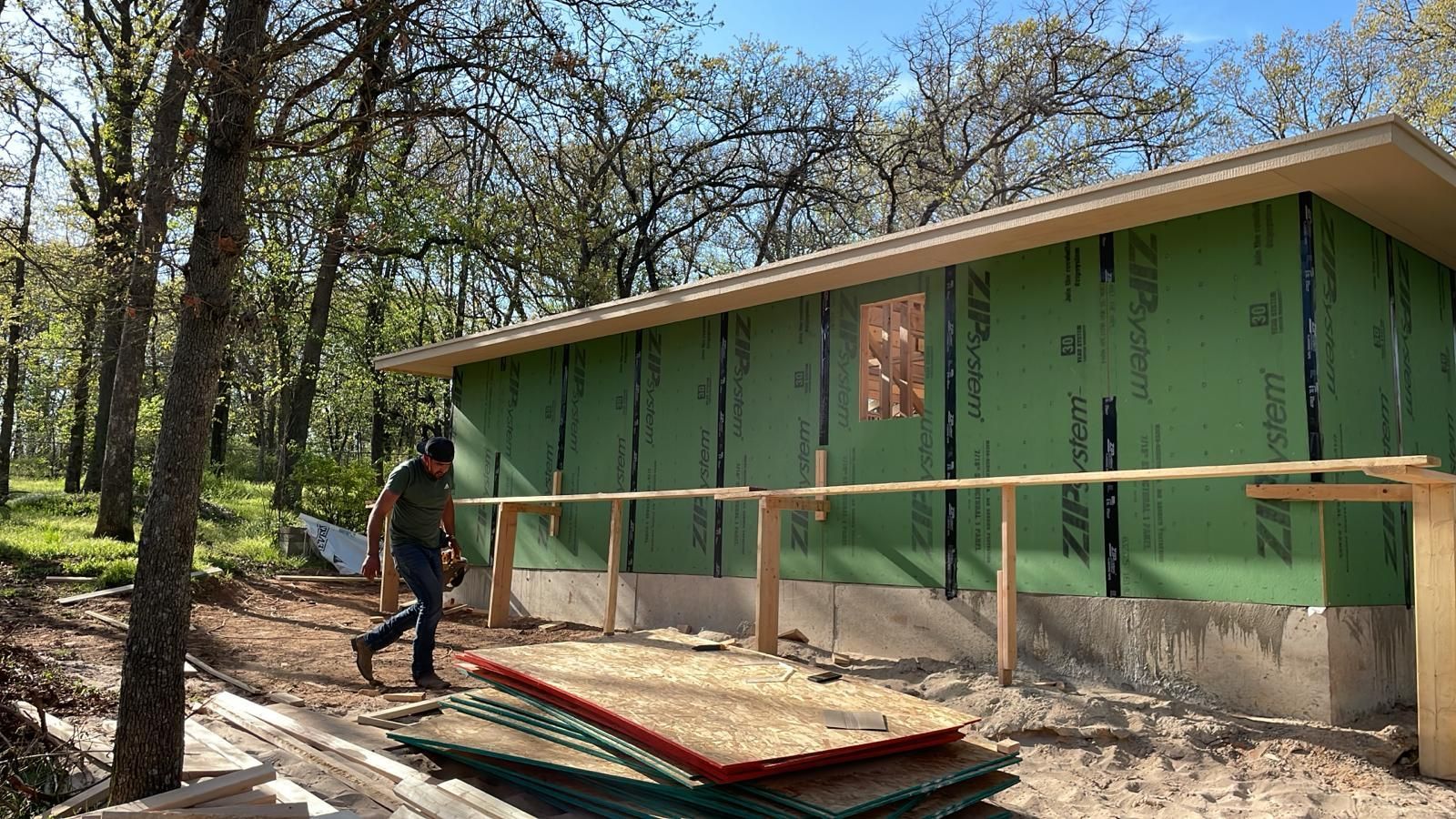 Man walking near a partially built building with green siding and a low wooden fence. Outdoors, daytime.