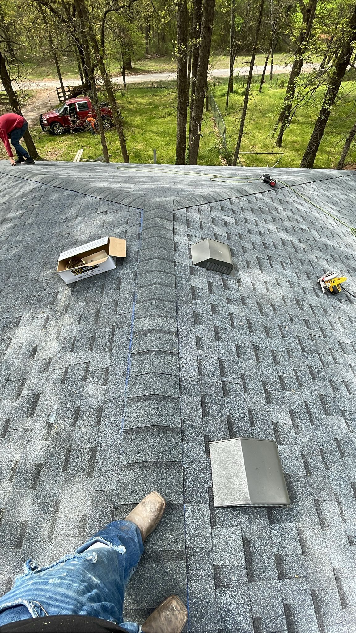 Person on a roof with asphalt shingles, supplies, and trees in the background.
