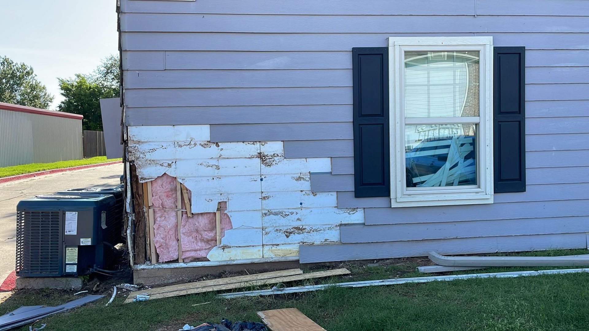 Damaged house siding next to a window; insulation and air conditioning units are visible.