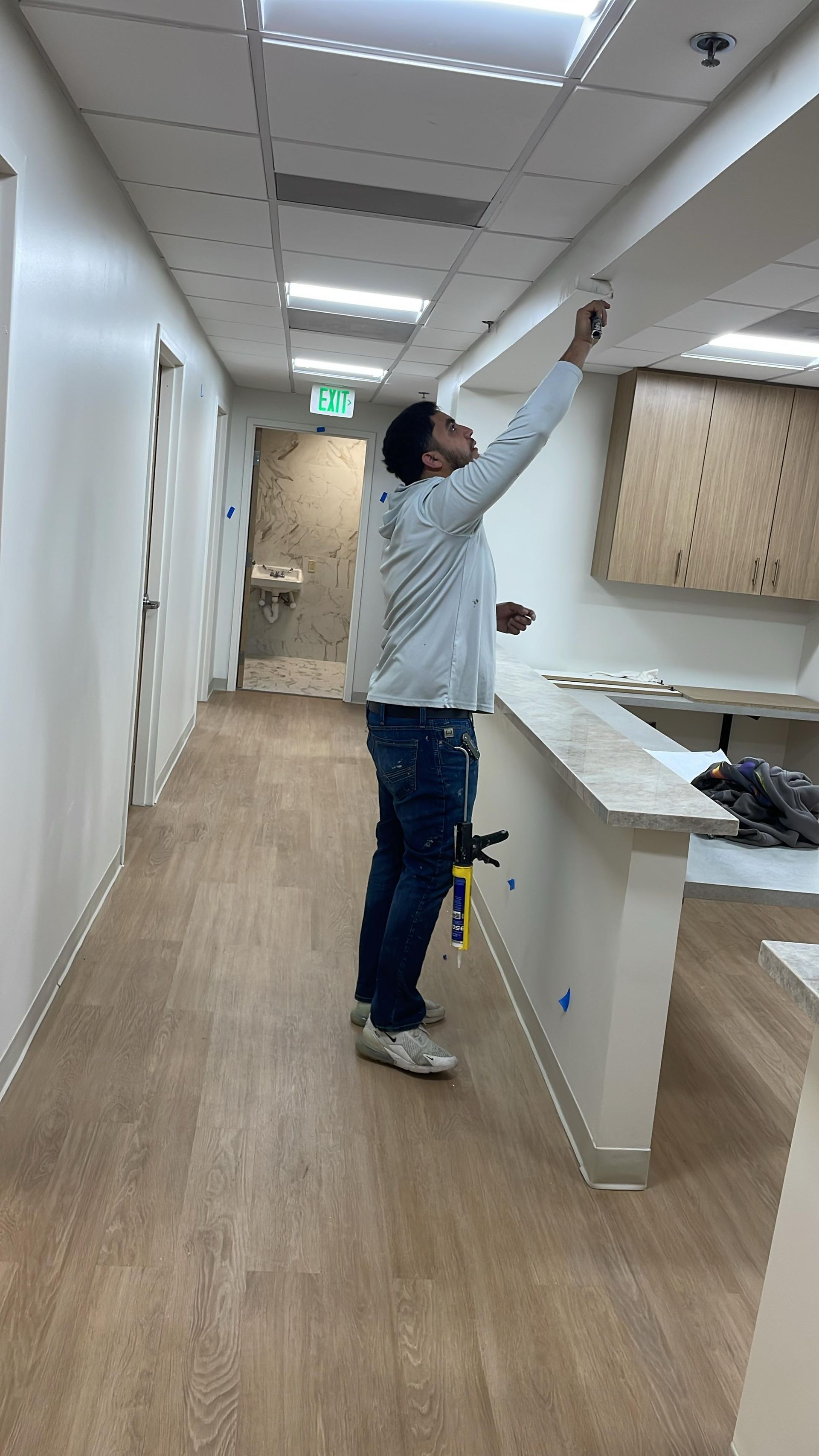 Person in a hallway reaching towards a ceiling fixture; beige walls and light wood flooring.