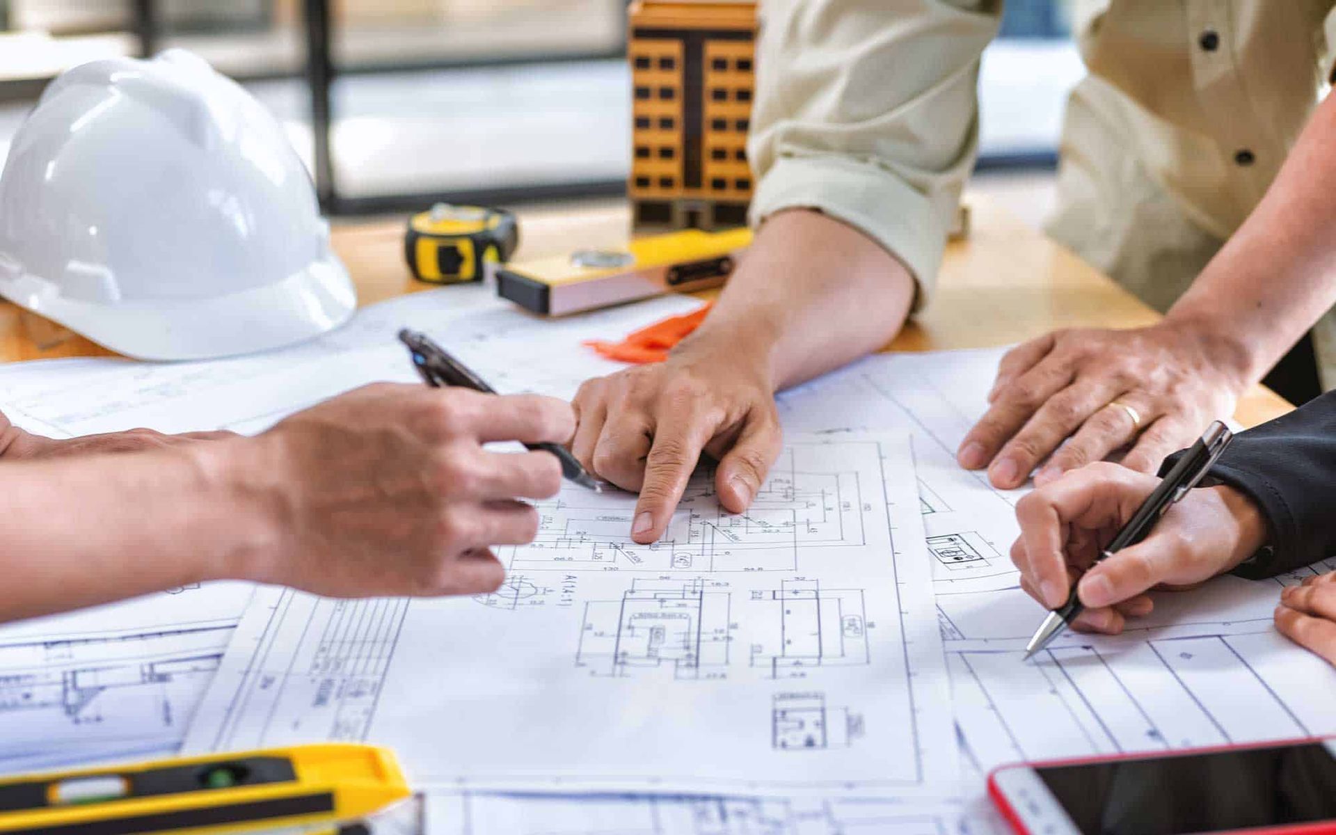 Construction workers review blueprints, pointing and writing. Table with tools and model building.