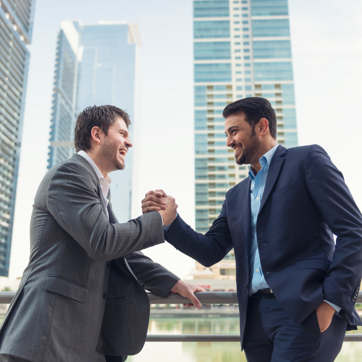 Two men in suits are shaking hands in front of tall buildings