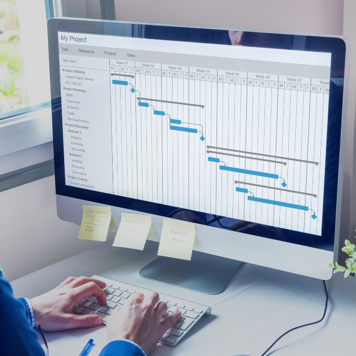 A person is typing on a keyboard in front of a computer monitor showing a project plan