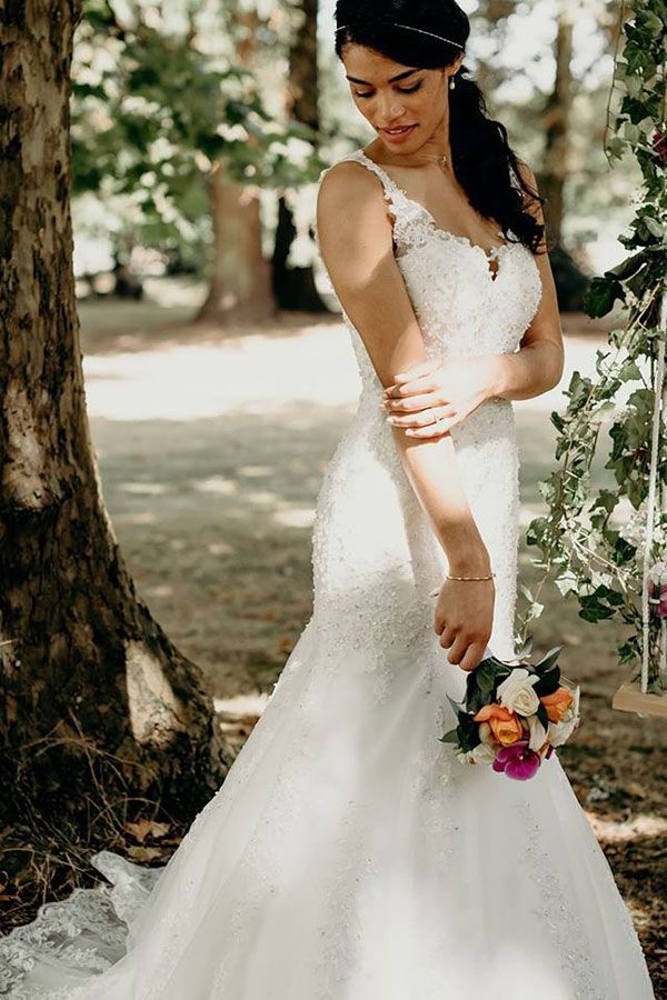 Une femme en robe de mariée se tient à côté d'un arbre tenant un bouquet de fleurs.