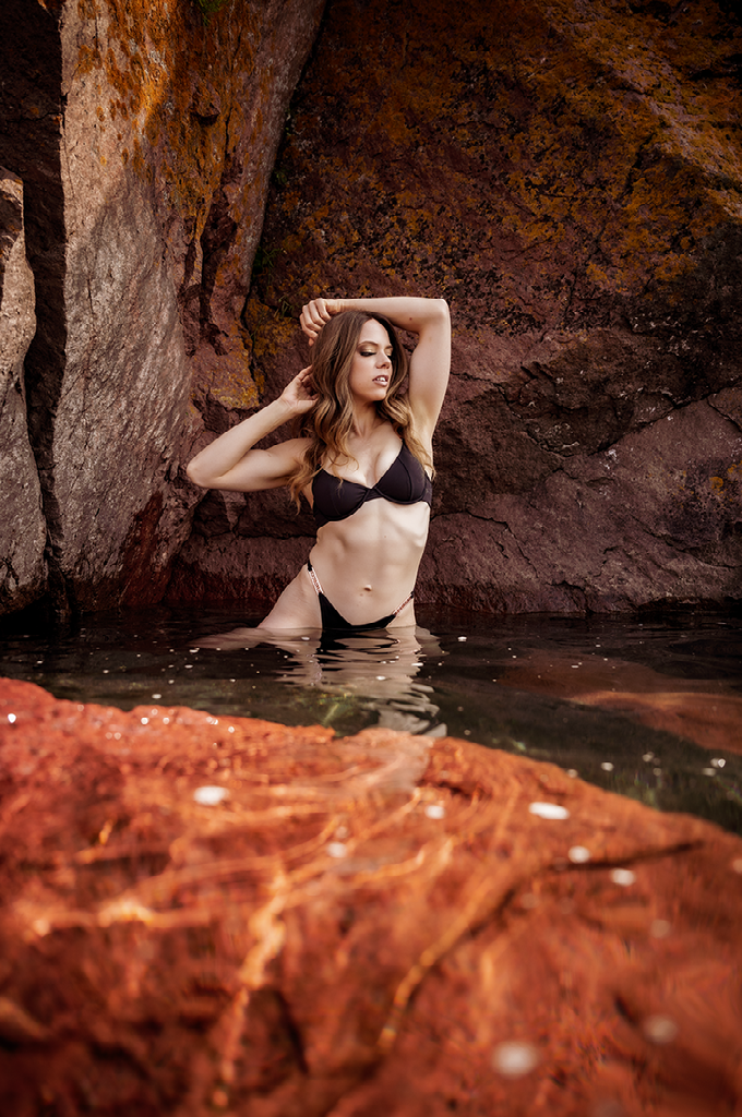 A woman in a bikini is standing in the water near a rock.