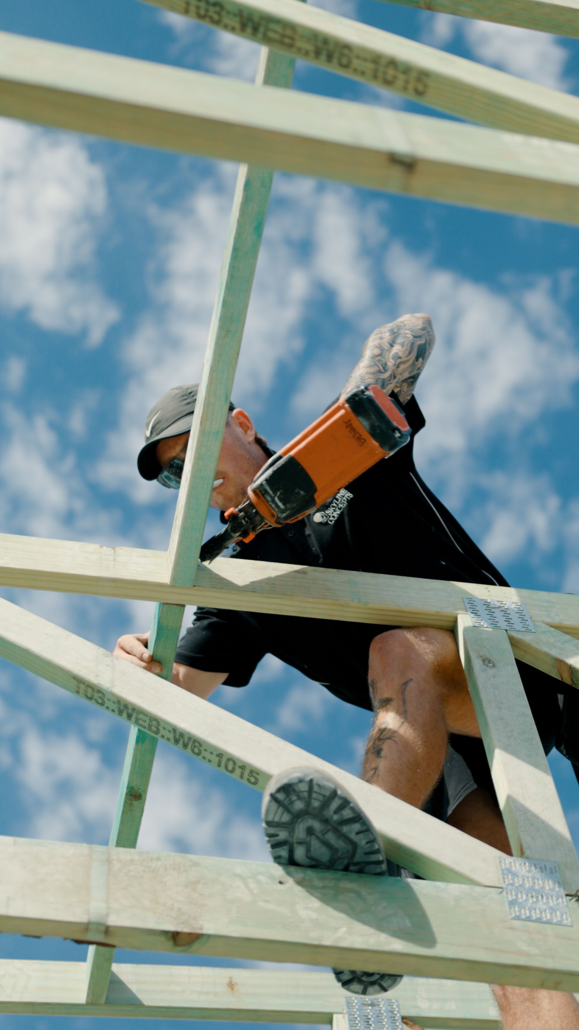 Person climbing a white railing outdoors under a blue sky, wearing black clothes and orange gear