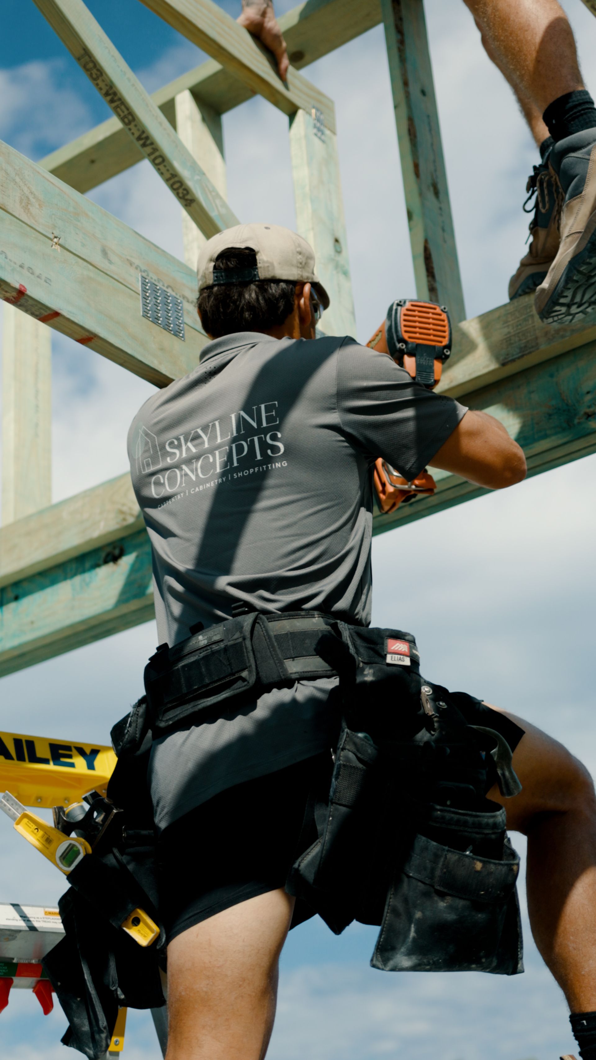 Worker in gray shirt and tool belt fastening beams on a wooden frame outdoors, with another worker above.