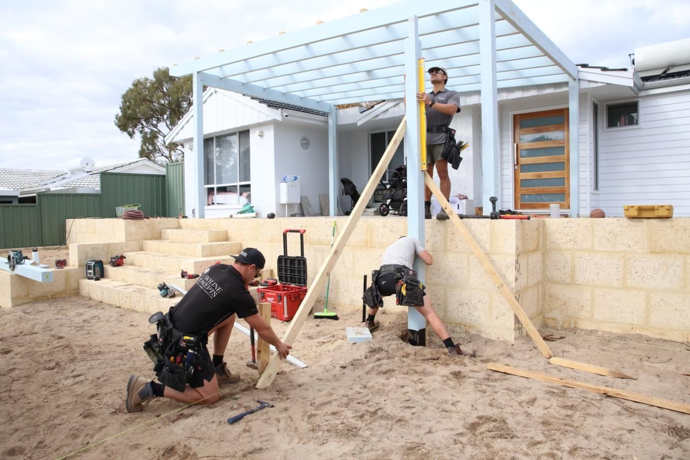 Workers building a backyard deck frame beside a white house, with tools and sand in the foreground