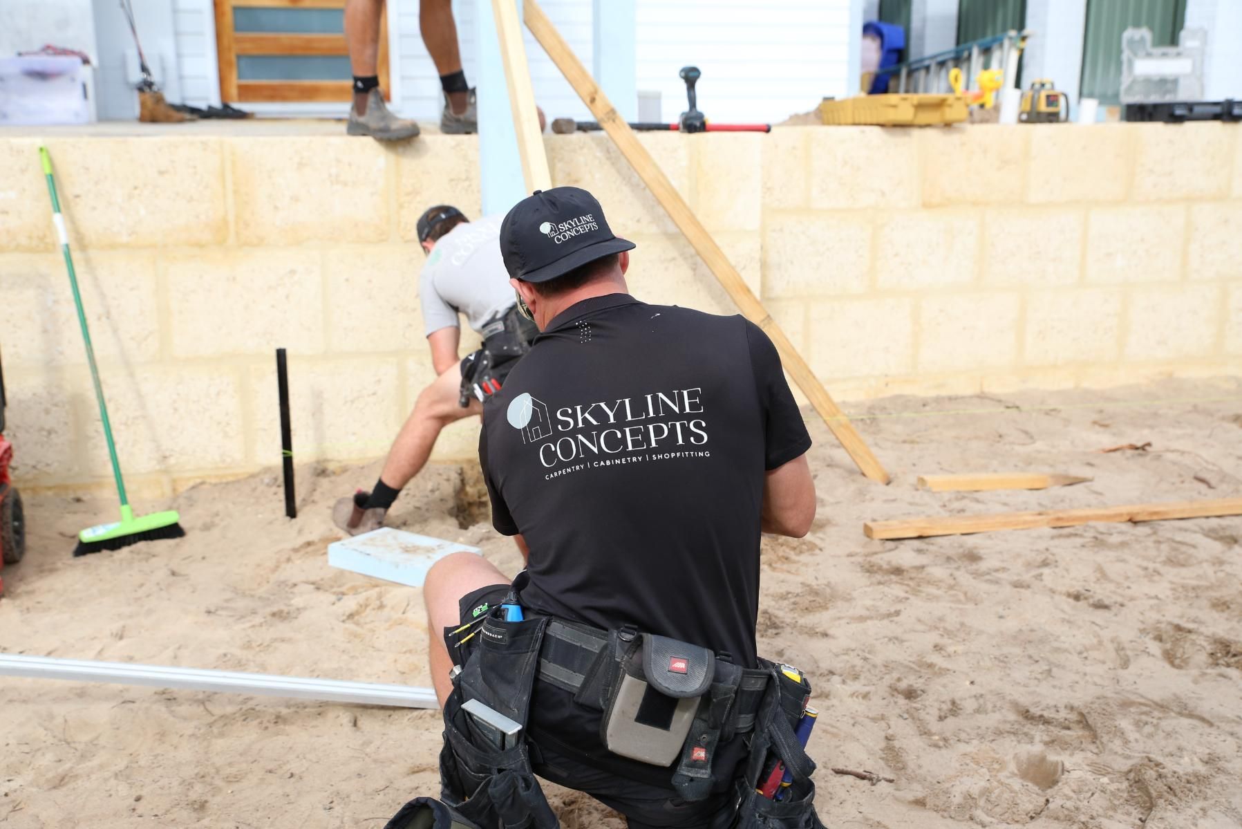 Two workers kneel at a sandy construction site beside a yellow block wall and stairs.