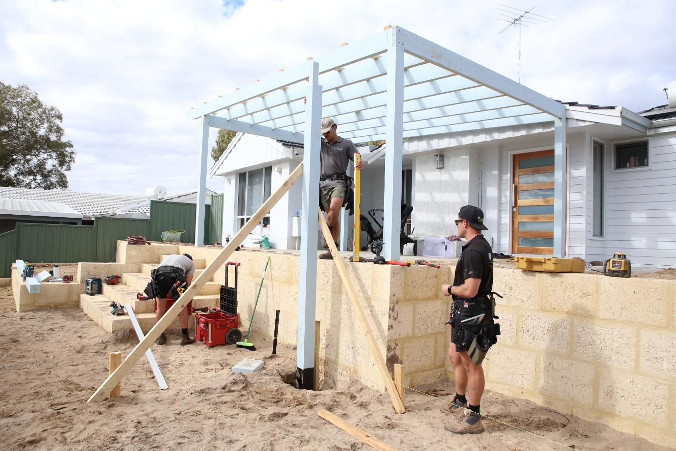 Workers building a patio frame beside a house, with sand, tools, and a partially constructed wall.