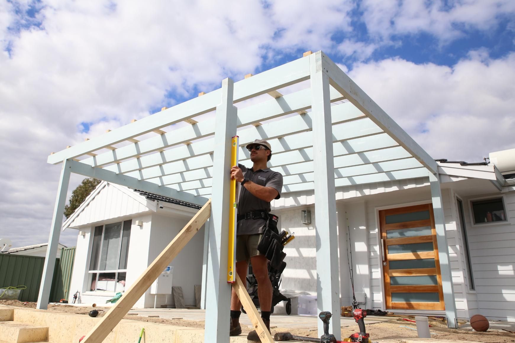 Worker standing on a ladder framing a white patio pergola outside a house under a cloudy sky