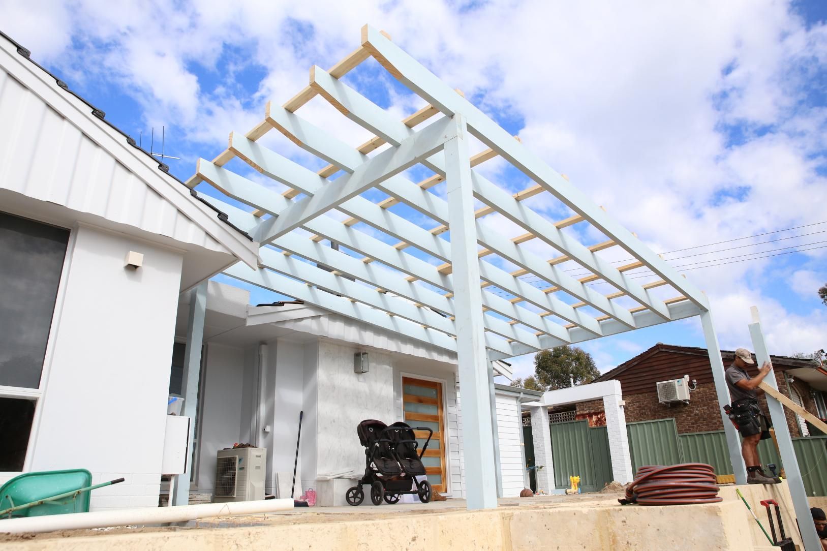 Backyard patio with a white pergola frame under construction beside a house on a sunny day