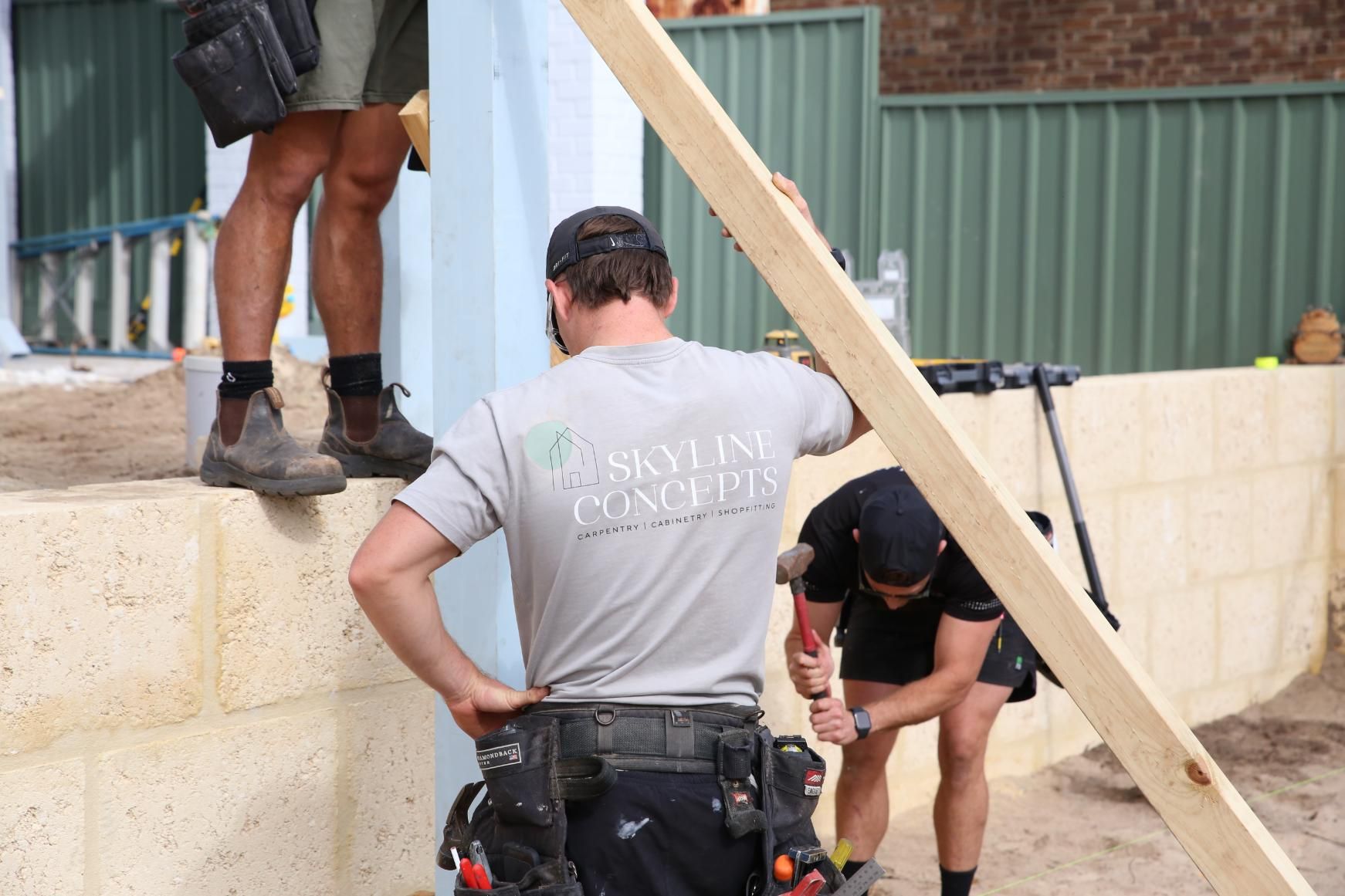 Workers installing a wooden beam on a construction site beside a corrugated metal wall.
