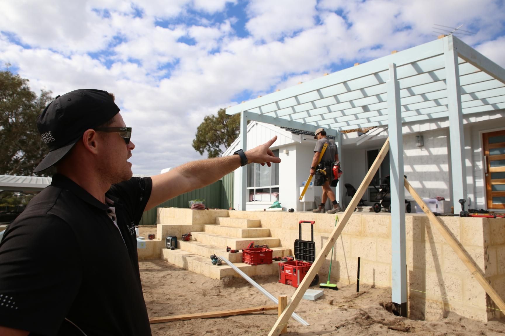 Man in black cap points at a house under construction with workers and exposed framing