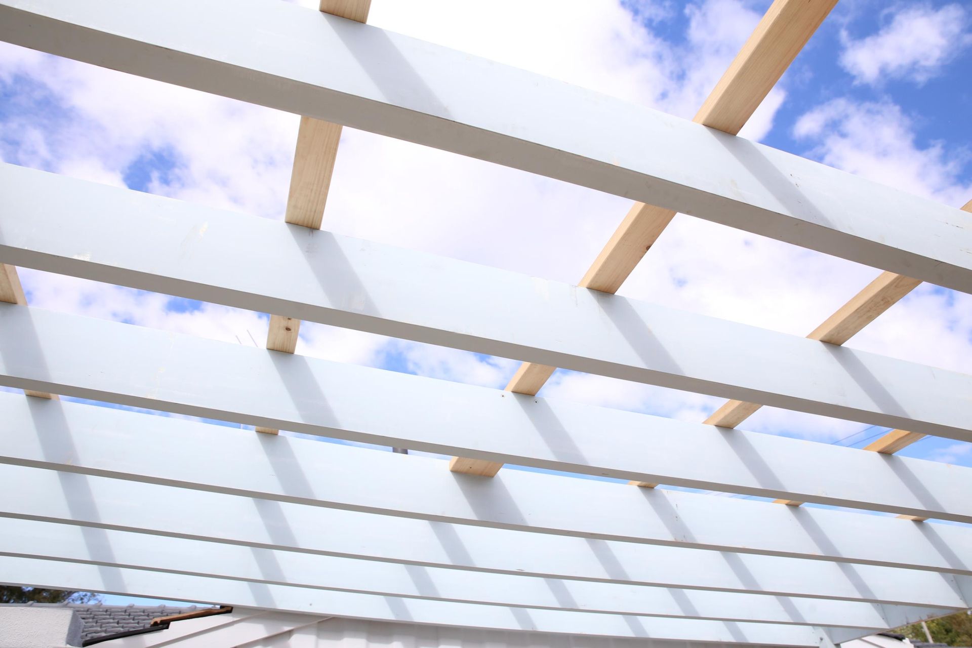 White pergola beams against a blue sky with clouds, viewed from below